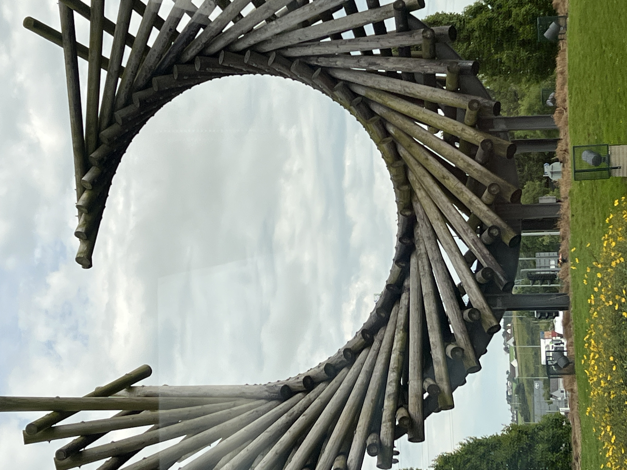 The image shows a close-up, abstract view of a wooden sculpture that appears to be part of a larger installation. The sculpture is made of many individual wooden poles or logs, arranged in a spiraling, circular pattern. The wood is weathered and appears to be a dark gray or brown color. The sculpture is viewed from below, with the sky visible through the open center. The sky is overcast with some diffused sunlight.
In the background, a grassy area with some yellow wildflowers is visible on the right side of the frame. Beyond the grass, there is a line of trees, and further in the distance, houses and buildings can be seen, suggesting an outdoor, possibly park-like, setting. There are also metal structures and some fencing visible in the background. The overall impression is one of an outdoor art installation in a natural or semi-urban environment. The time of day appears to be daytime, with cloudy weather conditions. There is no discernible activity or event taking place, and no people are visible. No text is visible in the image. The location context provided is Letterkenny, Ireland.
