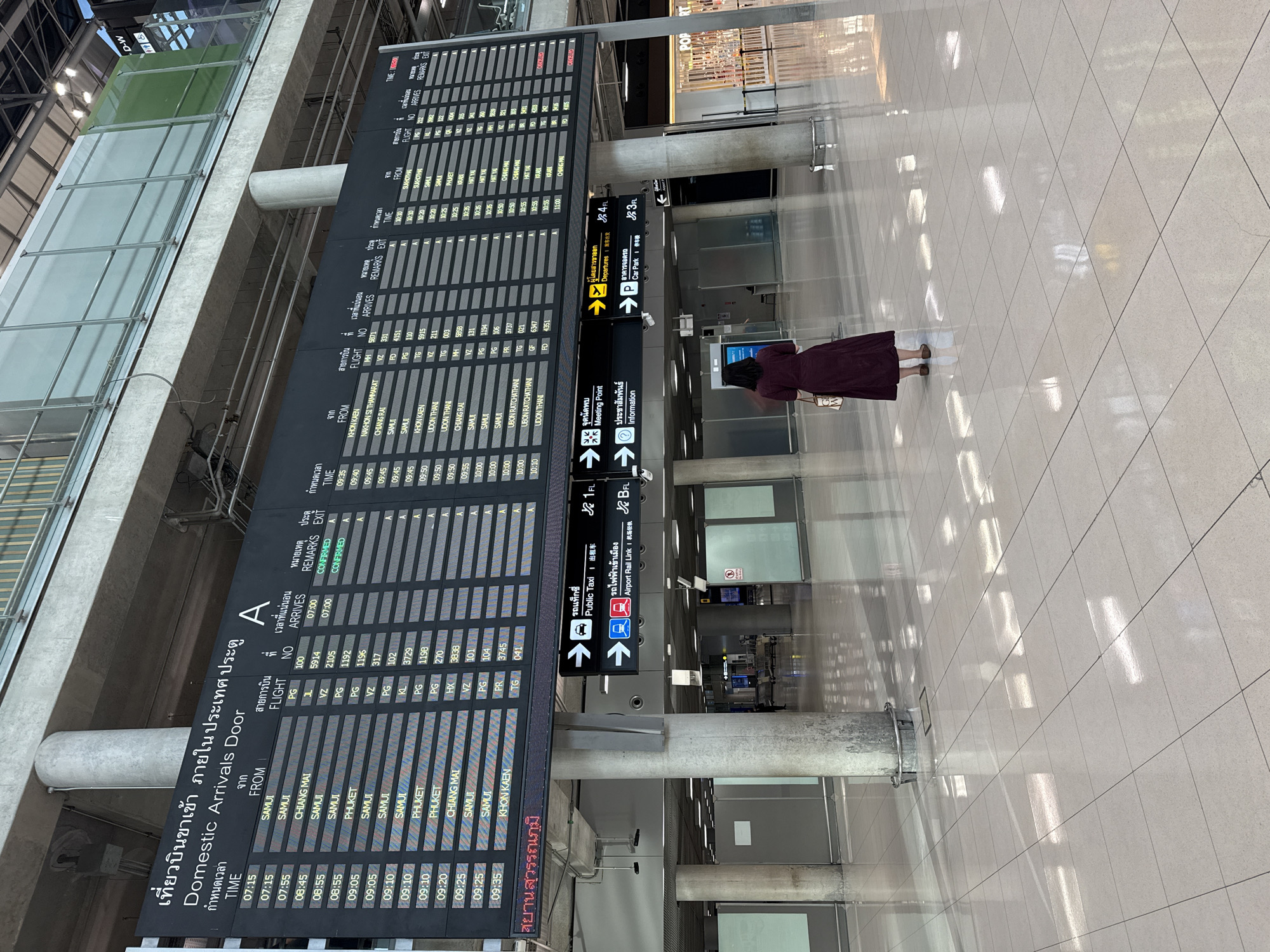 The image shows an interior view of an airport terminal. A large digital display board prominently features flight information for domestic arrivals. The board is divided into columns showing TIME, FROM, FLIGHT NO, ARRIVES, REMARKS, and EXIT. The text on the board is in both Thai and English. Below the flight information board, there are directional signs indicating Public Taxi, Airport Rail Link, Meeting Point, Information, Departures, and Car Park. A solitary person, a woman wearing a dark maroon dress and carrying a tote bag, stands in the foreground facing away from the camera, looking towards the display boards and signage. The environment appears to be a clean, modern airport concourse with a polished tiled floor and large concrete pillars. The lighting suggests it is daytime, with natural light filtering in and artificial lights illuminating the space.