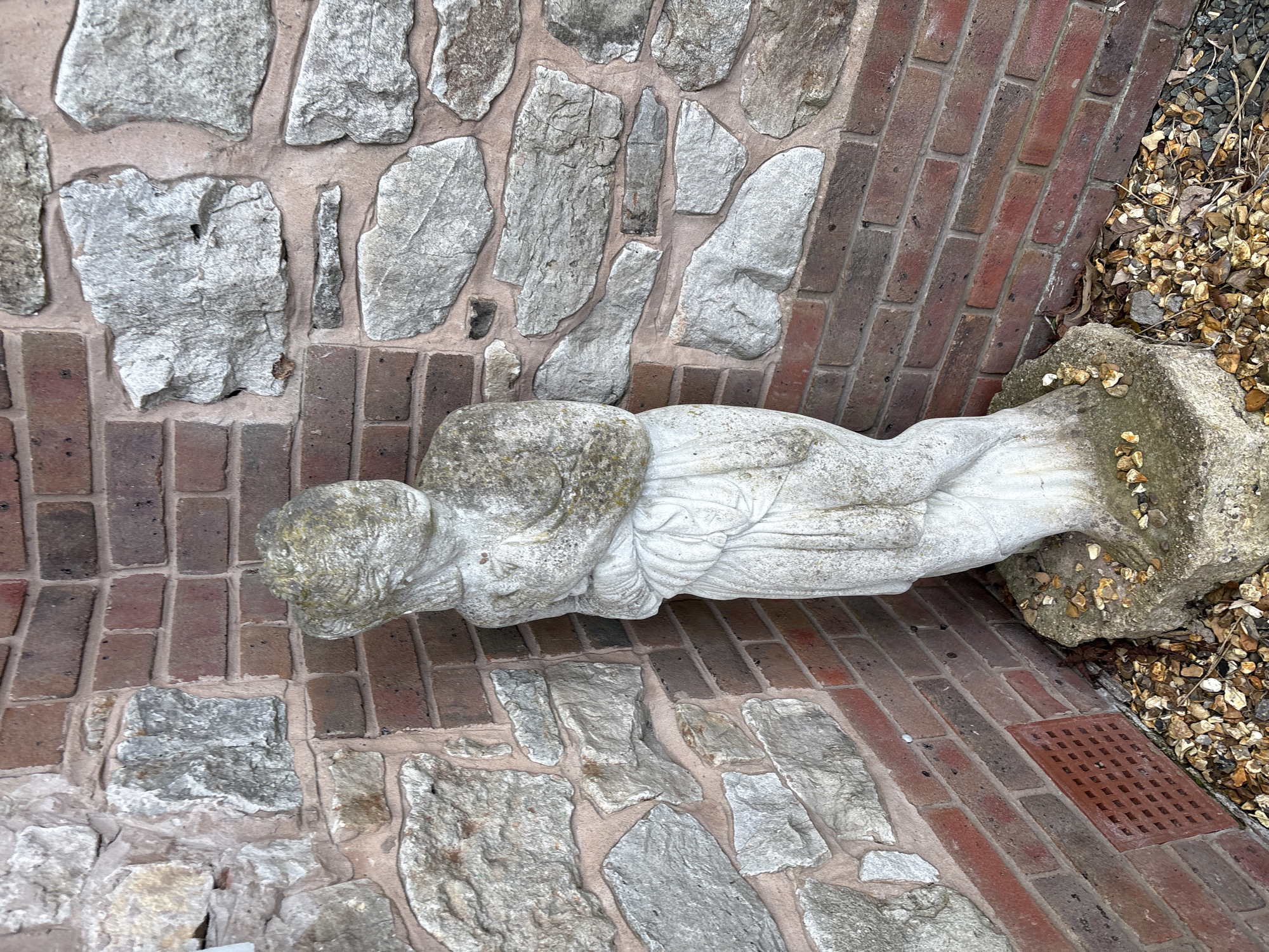 A pale stone statue of a reclining figure, likely an angel or similar allegorical figure, rests on a walkway constructed of red bricks alternating with sections of irregularly shaped grey stones. A stone plinth supporting the figure's lower body is surrounded by small pebbles and debris. The location appears to be outdoors, possibly in a garden or courtyard setting within the city of Shrewsbury, United Kingdom. A small drainage grate is visible near the base of the plinth.