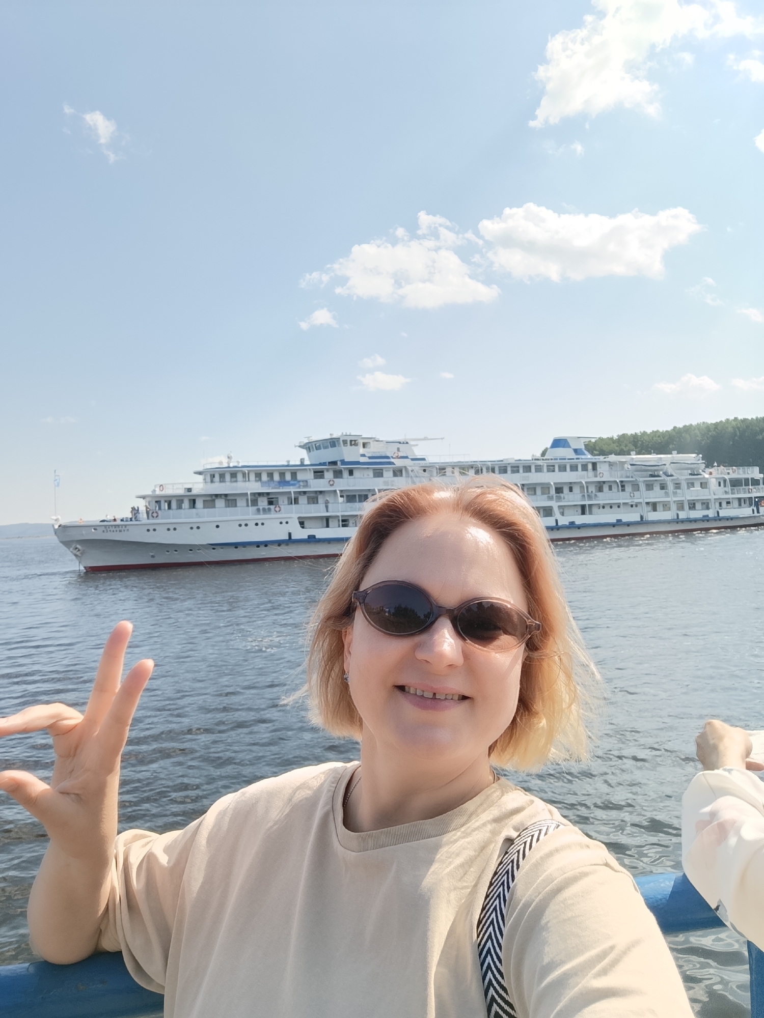 A selfie of a woman with blonde hair and sunglasses is taken in front of a large white passenger ship on a sunny day. She is wearing a beige t-shirt and has her left hand raised with two fingers extended in a peace sign. The ship, named "KAMA" on its bow, is docked or moving slowly on a body of water, possibly the Kama River, as indicated by the location of Chaikovsky, Russia, a city on the Kama River. The sky is blue with scattered white clouds. The woman's face is partially visible, and she is smiling. A blue railing is in the foreground.