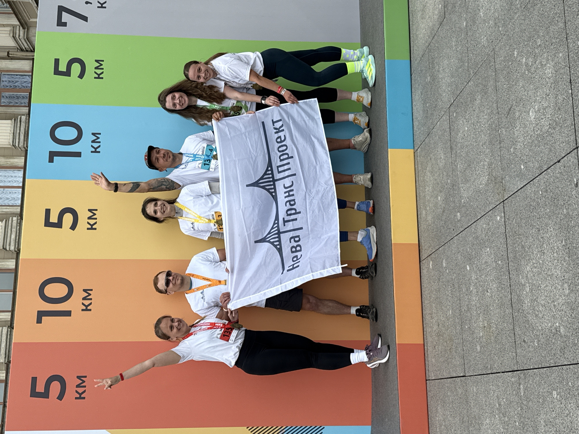 Six people, four women and two men, are standing on a raised platform in front of a brightly colored wall with large numbers indicating kilometers for a race. They are posing for a photo while holding a white flag with a bridge graphic and Russian text that reads "Нева Транс Проект" (Neva Trans Project). The people are wearing athletic clothing and race bibs. The man on the far left is wearing sunglasses. The wall behind them is divided into vertical sections of red, orange, yellow, light blue, green, and gray, with the numbers 5, 10, 5, and 7. The race appears to be a running event, likely a marathon, with distances of 5 km and 10 km advertised. The buildings visible in the background suggest an urban setting, and the architectural style is consistent with Saint Petersburg, Russia. The ground in the foreground is paved with large grey stone slabs.