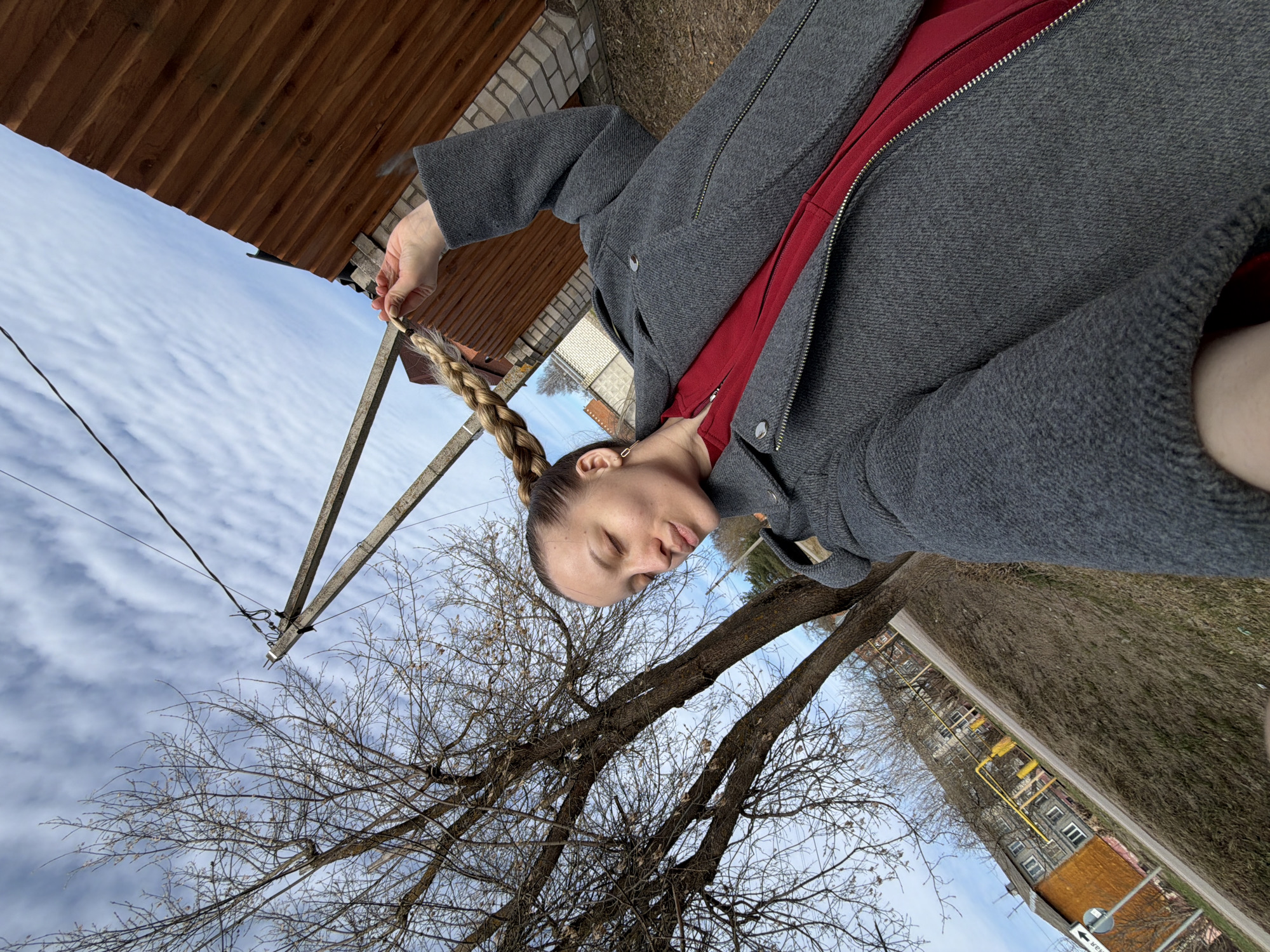 A person is standing outside in Reshetnikovo, Russia. The sky is visible with clouds and powerlines. The person is holding a braid of hair. A gray coat is zipped with a red garment showing underneath. A tree with bare branches is next to the person and across the street is a multi-story building.