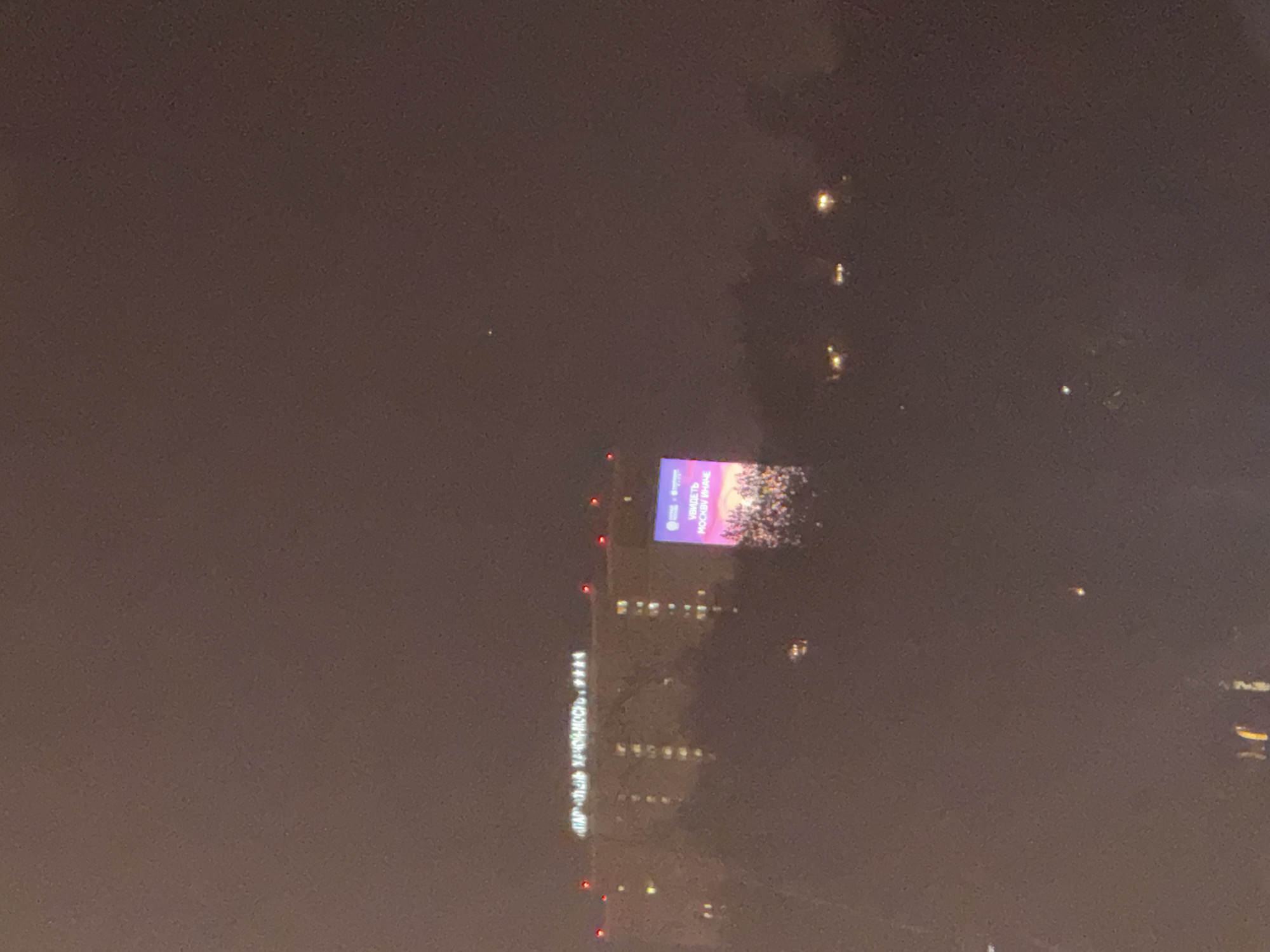The image depicts a nighttime view in the city of Mytishchi, Russia. The prominent feature is a building illuminated with lights, possibly a hotel or apartment complex, displaying the sign "ПАРК-ОТЕЛЬ ХВОЙНЫЙ." A large LED screen on the building shows an advertisement in Russian featuring a crowd and text that includes "УВИДЕТЬ МОСКВУ ИНАЧЕ." The scene is shrouded in fog or low visibility conditions, with scattered lights visible in the distance.