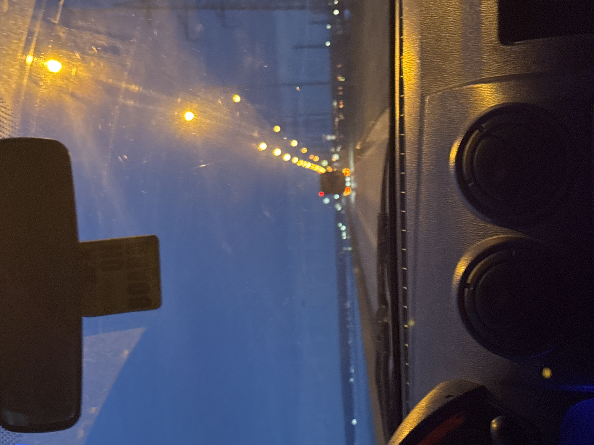 The image is taken from inside a vehicle, likely a car or truck, driving on a road at night. The view is through the windshield, which appears slightly dirty. A rearview mirror is visible on the left. The road is lined with streetlights, and other vehicles are present, with red taillights visible in the distance. The dashboard of the vehicle is visible on the right, including air vents. The location is identified as Noviy, Russia based on the location. No people are directly visible in the image.