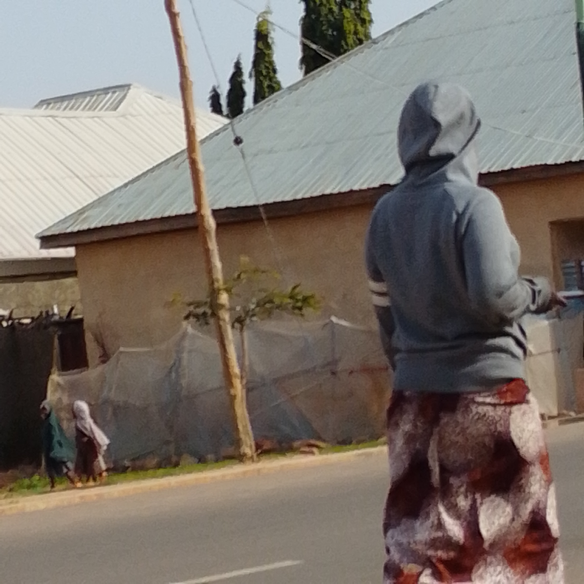 This image, taken in the city of Gombe, Nigeria, depicts a street scene with two individuals walking along the side of the road and another person standing facing away from the camera. The foreground shows a paved road. In the middle ground, two figures are walking from left to right. The figure on the left wears a dark garment, possibly a hijab, and the figure to its right wears a lighter colored hijab and a patterned skirt. They are walking alongside a low wall or structure. In the background, a person wearing a grey hooded sweatshirt and a long, patterned skirt stands facing away, with their back to the viewer. Behind them, there are buildings with corrugated metal roofs and stucco walls. Several tall, slender trees are visible behind the buildings. A wooden pole, possibly part of a utility line or fence, stands vertically in the left portion of the frame, obscuring part of the scene. The sky is clear and appears to be daytime.
