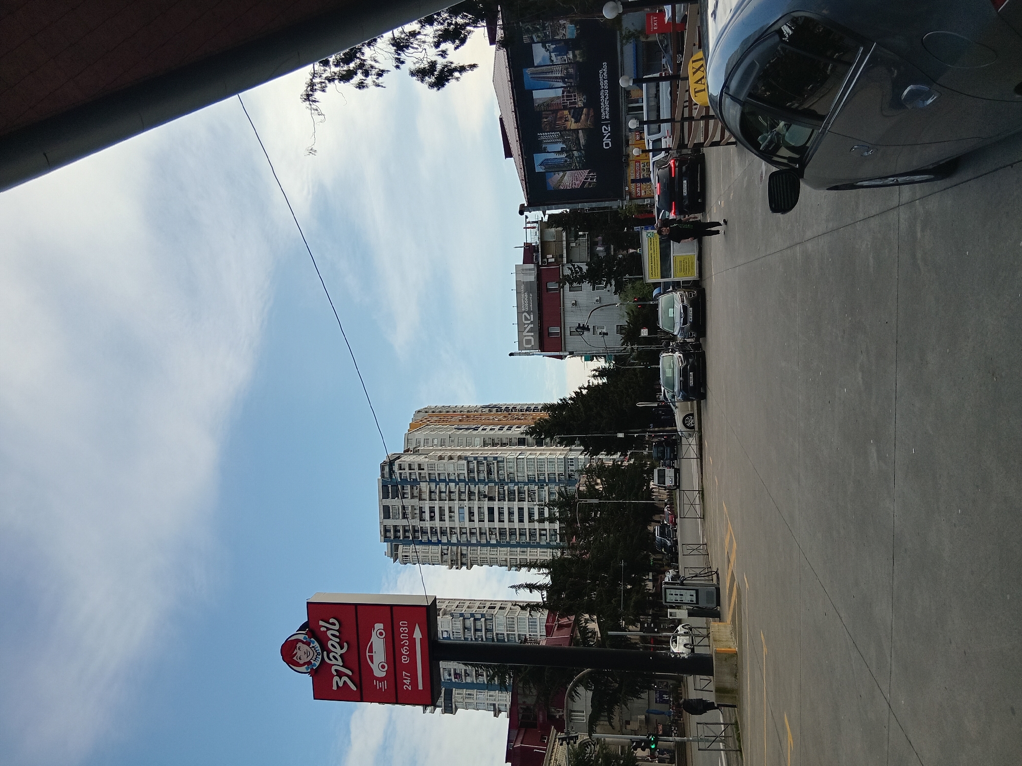 The image depicts an urban environment in Batumi, Georgia. The sky is light blue with scattered clouds. Several high-rise buildings are visible, including one with a blue and white facade. A red and white sign is prominently displayed, featuring a depiction of a car and Georgian text. The location appears to be near a parking area or street with vehicles and a taxi stand. A person is walking on the sidewalk. Streetlights, advertisements and utility poles are also present.