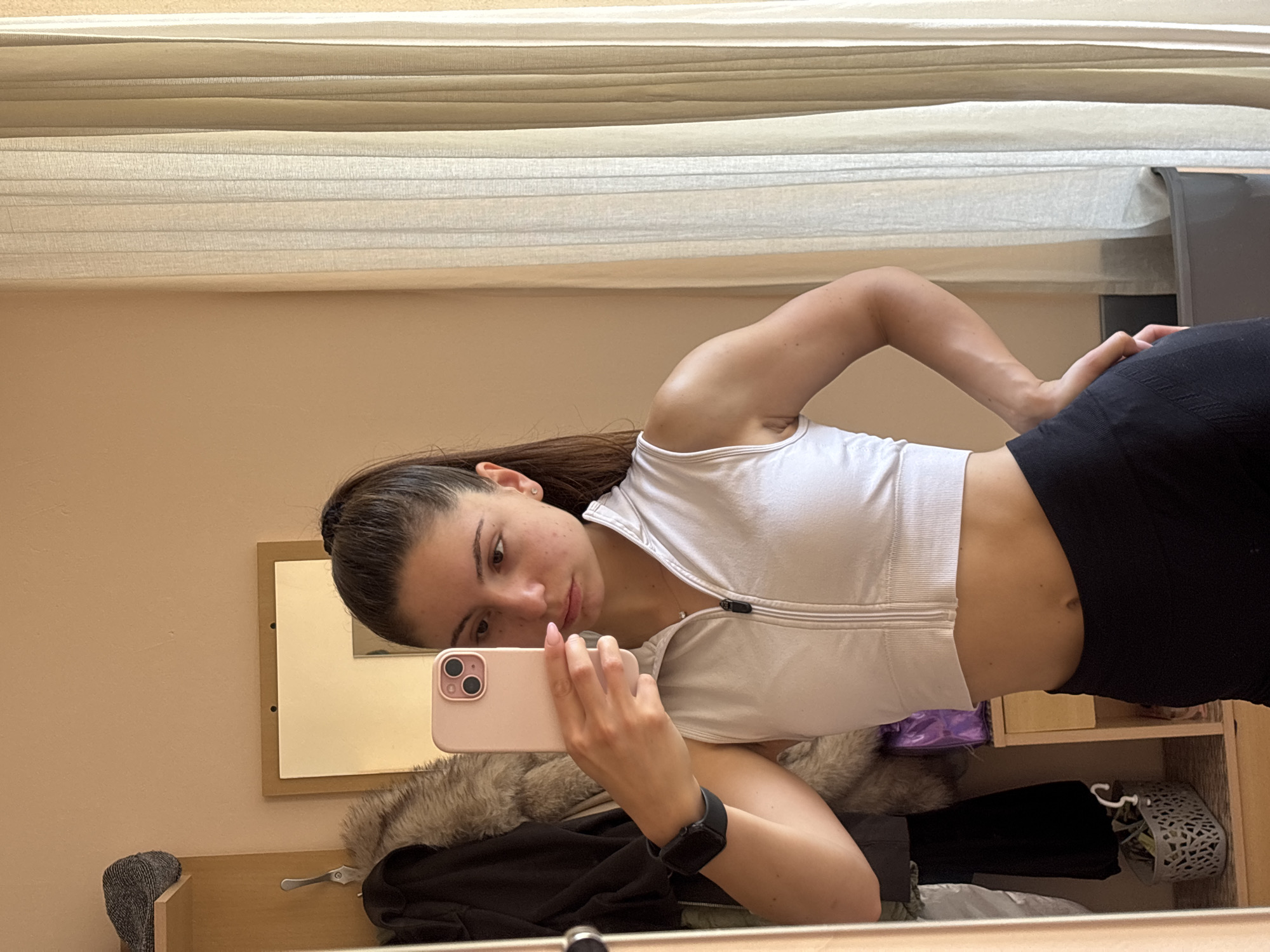 A young woman with dark hair tied in a high ponytail is depicted taking a mirror selfie indoors, in what is stated to be a location in Varna, Bulgaria. She is positioned facing the mirror, holding a pink smartphone in her right hand to capture her reflection. Her left hand rests on her left hip. She wears a white, sleeveless, collared athletic top with a black zipper extending from the neckline to the mid-chest, revealing her midriff. Her bottom garment consists of black, high-waisted athletic leggings. A black smartwatch is visible on her left wrist, and a delicate silver necklace with a small pendant is around her neck. Her expression is neutral to slightly pouting, and she has visible blemishes on her face.
The immediate environment visible in the mirror's reflection includes a light beige or peach-colored wall on the left. To the left of the woman, a wooden shelving unit or part of a wardrobe is present. On the top shelf, a grey knitted hat is placed. Below, a silver hook holds two garments: a black jacket and another garment with a light brown fur collar. On a lower shelf, a white woven basket containing various small items is visible. Above the wardrobe/shelf, a rectangular mirror or framed picture with a white or light-colored surface is partially reflected.
To the right of the woman, a floor-length, light-colored curtain with vertical pleats covers a window or wall, extending from the top to the bottom of the frame. The wall behind the curtain appears to be a similar light beige hue. A dark grey or black object, possibly a storage bin, is partially visible at the bottom right, next to the curtain. The overall lighting suggests an indoor setting, likely with natural light filtering in from the right.