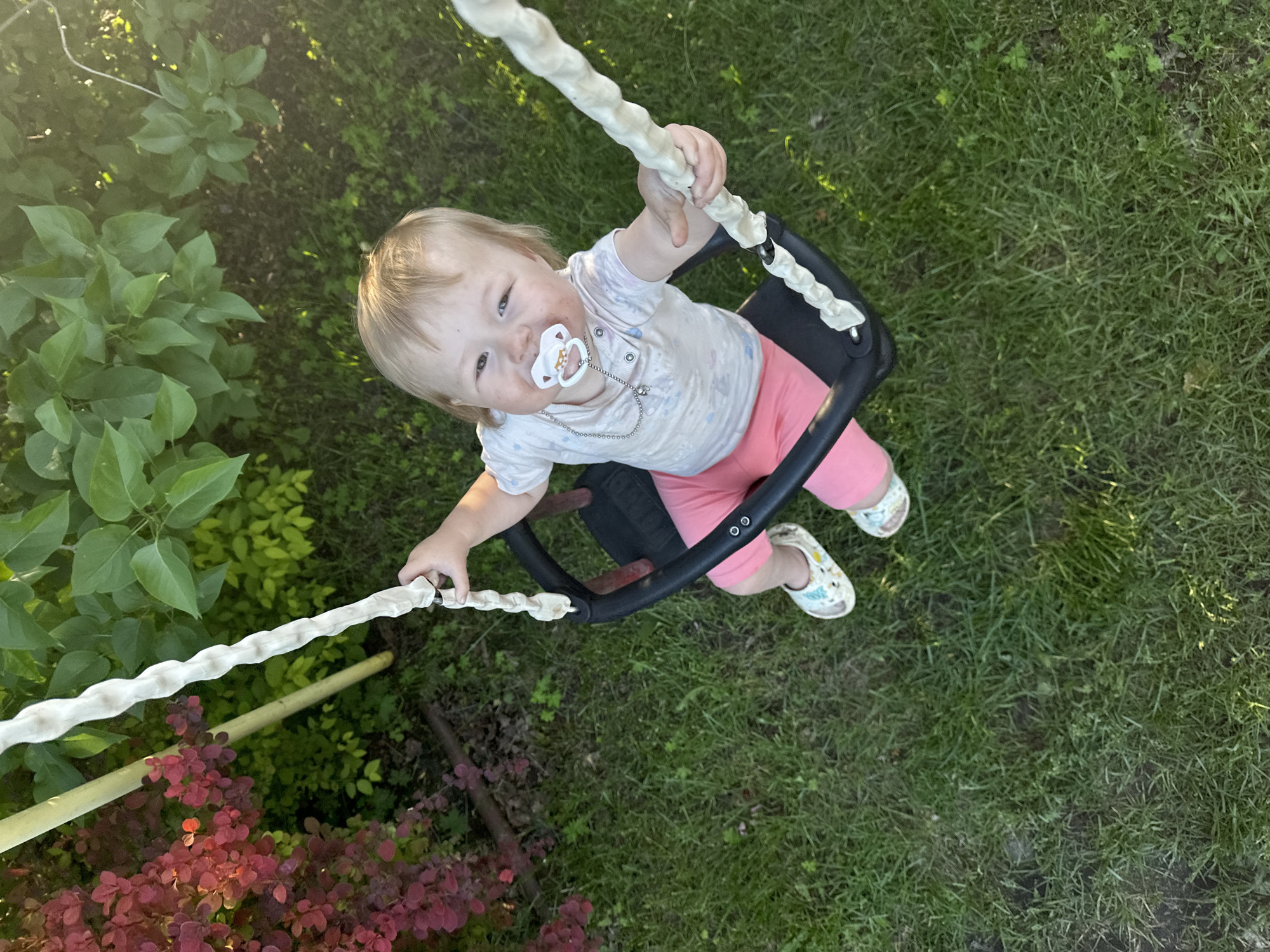 An outdoor scene depicts a young child seated in a black bucket swing, viewed from a high-angle perspective looking downwards. The child, a toddler with blonde hair, wears a light-colored short-sleeved top, pink shorts, and white sandals with colorful patterns. A white pacifier with a clear and brown design is positioned in the child's mouth, attached via a beaded chain. Smudges are present on the child's face around the mouth and cheeks. Both of the child's hands firmly grip the swing's support ropes, which are covered in a textured, light-colored, possibly fabric or padded material. The ground beneath the swing is covered with green grass. In the background, to the left, a dense bush with deep red foliage is visible, alongside a slender, light-colored pole. To the right and in the upper background, various types of green-leaved bushes and trees are present. Strong sunlight illuminates the scene from the upper right, casting highlights on the foliage and the child. The specific location within the city of Nizhny Novgorod, Russia, cannot be determined from the visual evidence provided.