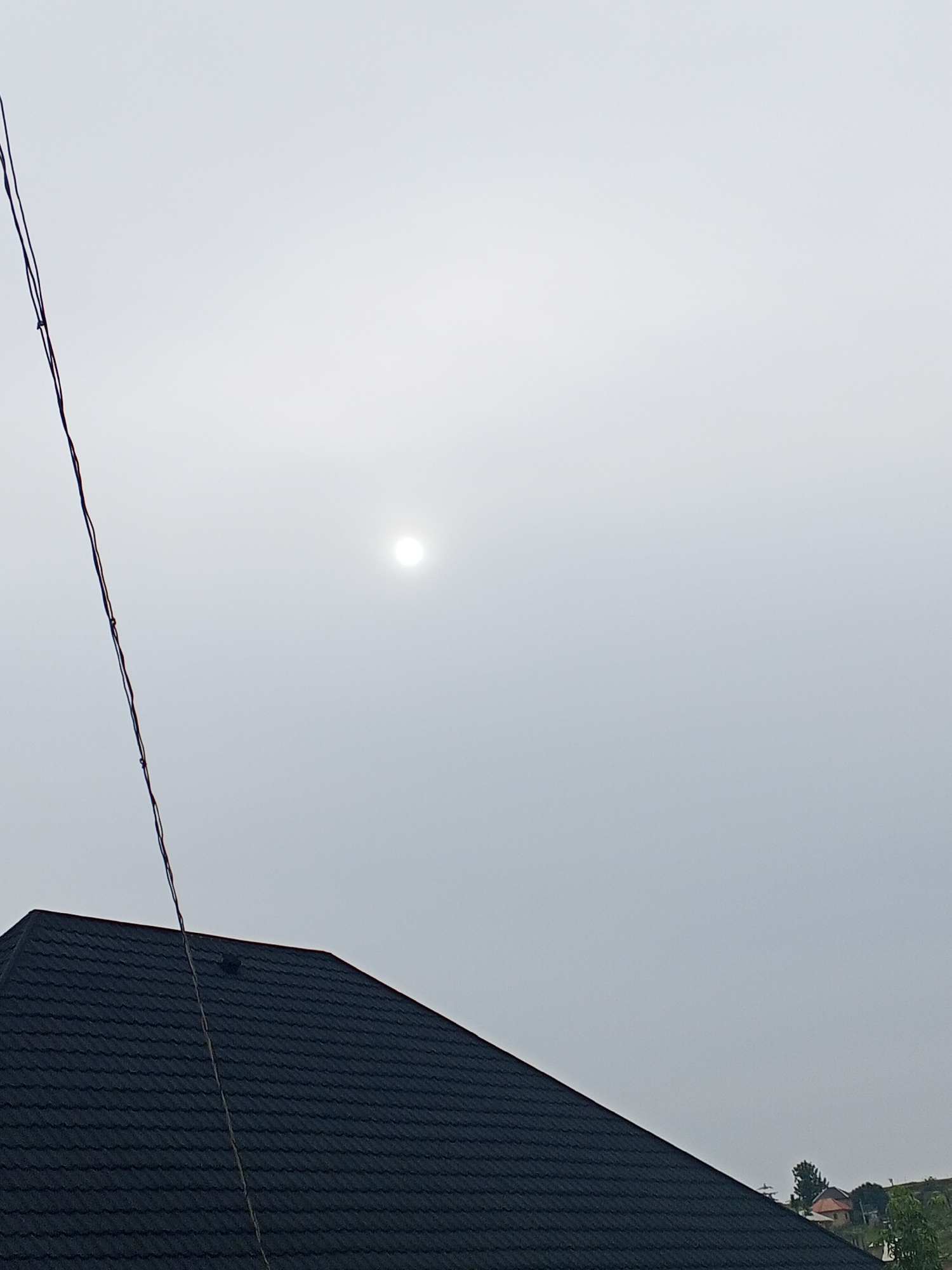 This is a full-frame, eye-level photograph taken outdoors in Ashata, Nigeria. The image features a dark, textured roof in the foreground, angled from the bottom left corner upwards towards the right. The roof appears to be made of asphalt shingles. A single, thin black wire runs diagonally from the top left edge of the frame down towards the roof. In the upper portion of the image, a bright, diffused sun is visible through a hazy, overcast sky. The sky occupies the majority of the frame, creating a muted and light-filled atmosphere. In the far background, on the right side of the frame, a few indistinct residential structures and some green foliage can be seen. There are no people or animals present in the scene. The overall impression is one of a cloudy day with the sun partially obscured.