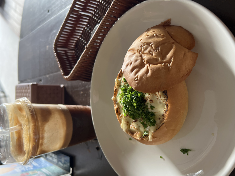 The image displays a meal set on a dark table surface in Nha Trang, Vietnam. A white ceramic plate centrally holds a round, light brown bread bowl filled with a baked, creamy substance garnished with finely chopped green herbs. The detached, sesame-seeded top of the bread bowl rests on the plate to the right of the main bowl. In the upper left quadrant, a clear plastic cup contains a layered iced beverage, featuring a dark brown liquid at the bottom and a lighter, foamy layer on top, with a visible straw. An empty, dark brown woven wicker basket is positioned to the upper right of the plate. A small, rectangular brown box is partially visible behind the beverage cup. Strong natural light illuminates the scene, creating distinct shadows on the plate. No individuals are present.