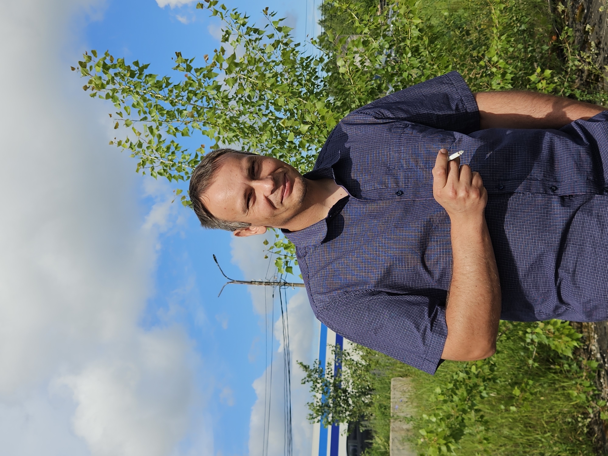 A man, appearing to be in his late 30s or early 40s, stands outdoors in front of a vibrant green tree and a bright blue sky with scattered clouds. He is wearing a short-sleeved, dark blue plaid shirt. In his right hand, he holds a lit cigarette between his fingers. The man is looking towards the camera with a slight smile. Behind him, partially visible through the greenery, is a building with a distinctive blue and white striped facade. A lamppost with electrical wires is also present in the background. The overall setting suggests an urban or industrial area, possibly in Chaikovsky, Russia, given the context. The ground in the foreground appears to be a mix of dark soil and gravel.