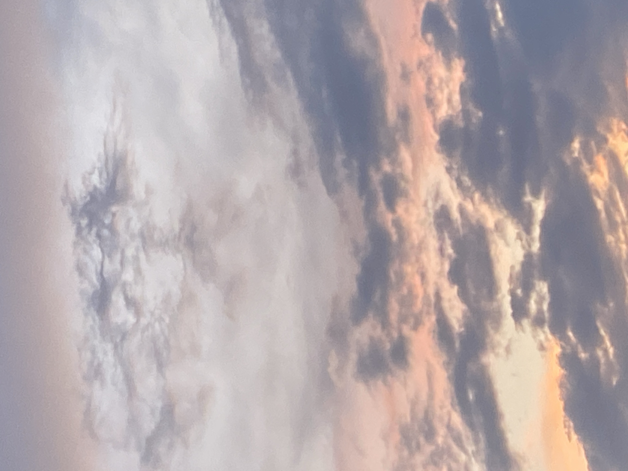The image shows a close-up, detailed view of a cloudy sky, likely during sunrise or sunset, as indicated by the soft, warm hues of pink and orange interspersed with gray and white clouds. The clouds are varied in texture and density, with some appearing wispy and translucent while others are thicker and more defined. The lighting suggests a transition period of the day, casting subtle shadows and highlights across the cloud formations. There are no people, objects, or specific landmarks visible that would indicate a particular location within Kushchovskaya, Russia. The scene is entirely focused on the atmospheric conditions of the sky.