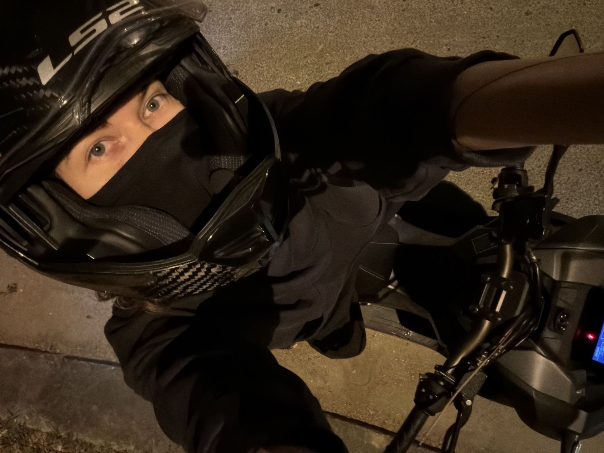 A person wearing a black helmet and black face mask is taking a selfie while sitting on a motorcycle. The helmet has the letters "LS2" on the front. The person's eyes are visible above the mask. The motorcycle handlebars and instrument panel are visible. The background appears to be a dimly lit street. The location is not possible to determine from the provided image.