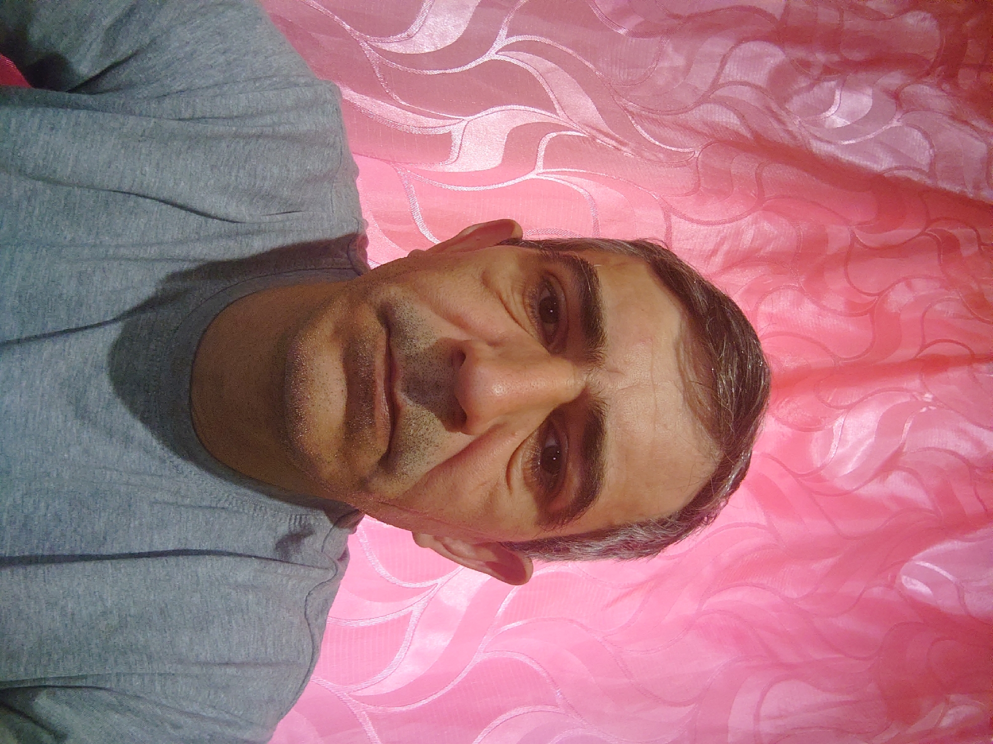 A middle-aged man with short graying hair and a faint stubble on his face is shown in a close-up, head-and-shoulders shot. He is wearing a gray t-shirt. The background consists of a pink curtain with a repeating pattern of stylized, swirling leaves. The man's expression is neutral. The lighting is even, casting minimal shadows. The image appears to be a selfie, taken indoors.