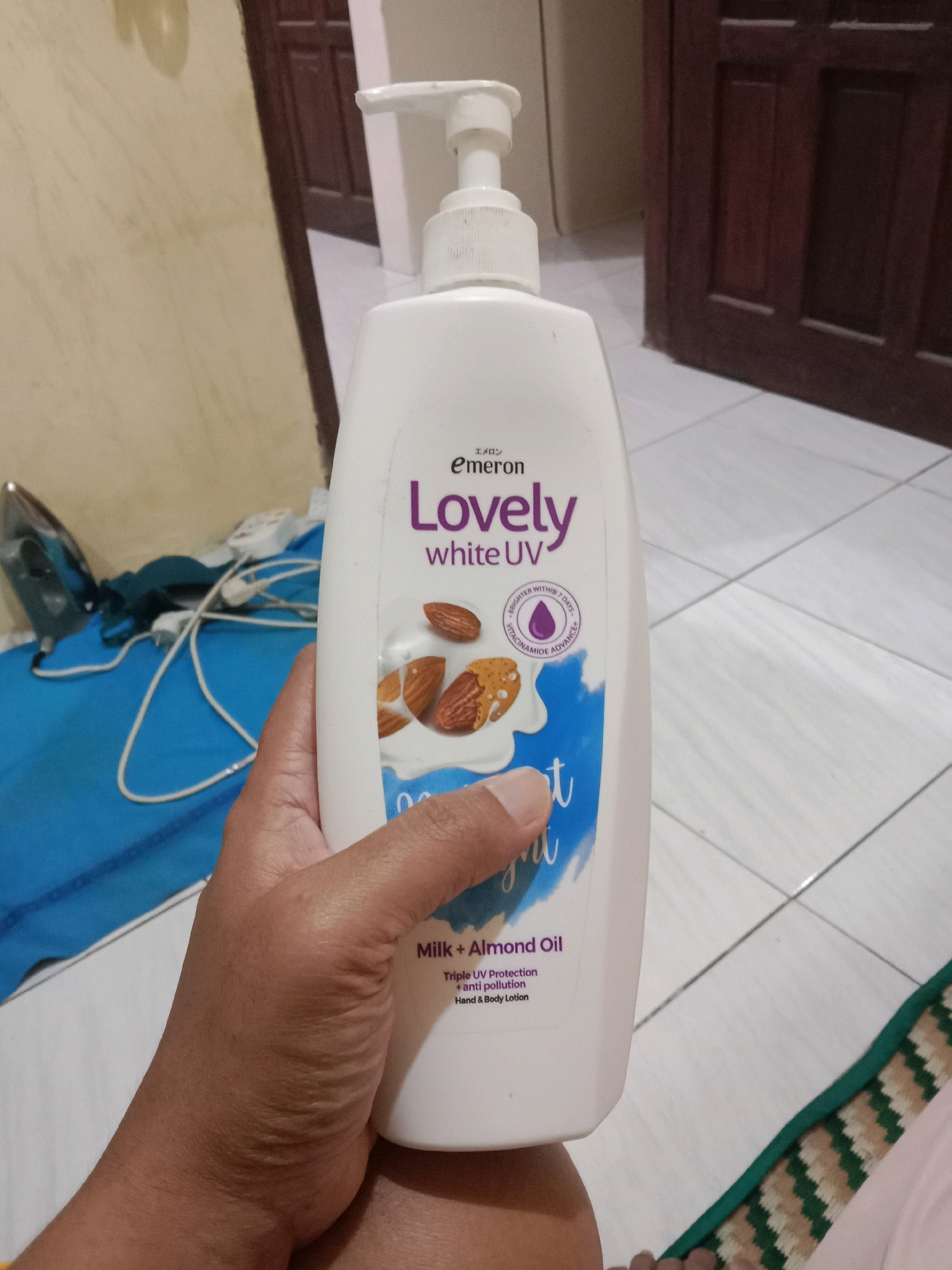 A person's hand holds a white pump bottle of "Emerson Lovely white UV Milk + Almond Oil" lotion. The bottle features images of almonds and milk. The background includes a tiled floor, a wall, and a wooden door. On the left side of the image, an iron and some blue fabric are visible. The scene appears to be indoors, possibly in a residential setting in Brangsong, Indonesia.