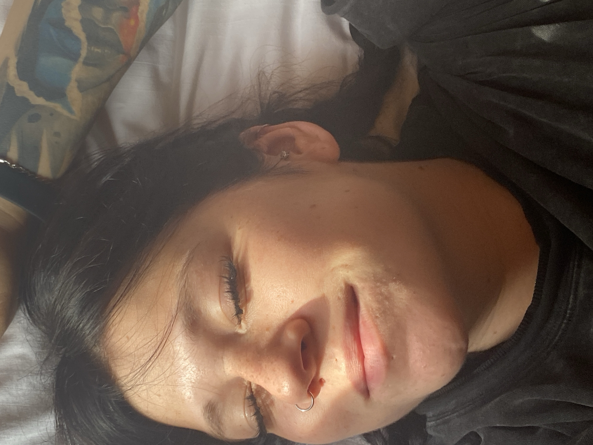 The image shows a close-up, low-angle shot of a young person lying down, with their eyes closed and a faint smile. Sunlight illuminates their face, highlighting freckles across their nose and cheeks. They are wearing a dark, presumably cotton, hooded sweatshirt. A silver hoop nose ring is visible in their left nostril, and a small silver stud earring is in their right ear. Their dark hair is partially visible, framing their face and extending upwards, where a tattooed arm is also visible. The background consists of a white, slightly textured surface, likely a bedsheet. The overall impression is one of peacefulness and relaxation, possibly captured in a bedroom setting. There is no visible indication of the city of Krasnoflotskoe, Russia.