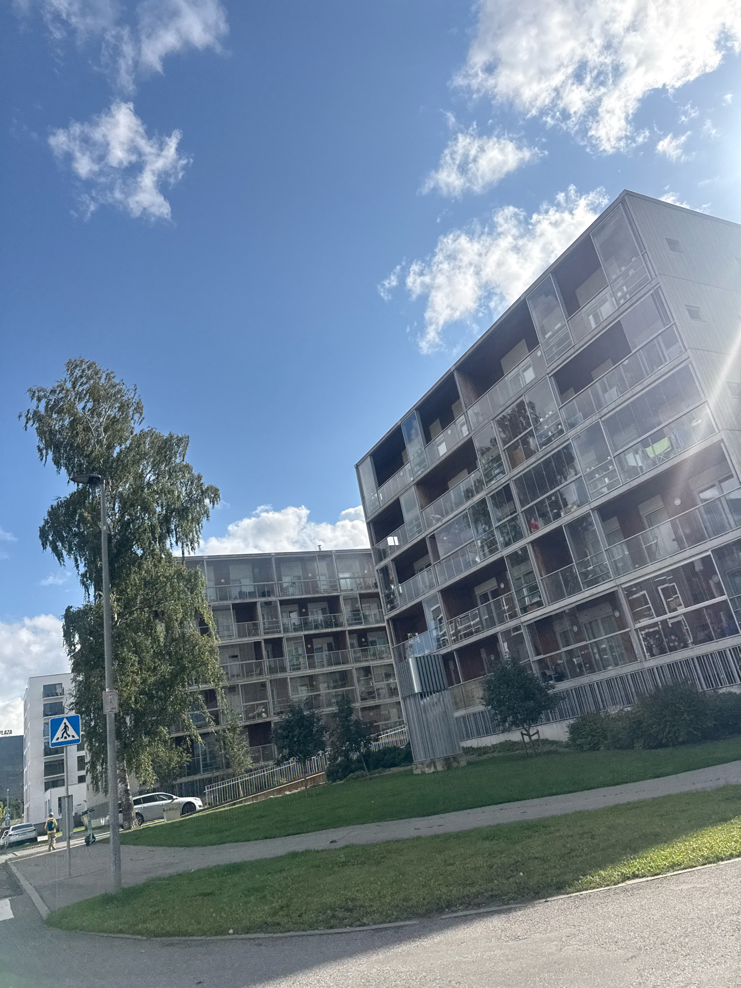 In Tallinn, Estonia, an image captures a contemporary apartment building with a glass facade, its balconies and windows reflecting the sky and surrounding trees. To the left, a slender, tall tree with sparse foliage stands next to a lamppost. A pedestrian crossing sign is visible on the sidewalk, with a person walking in the distance. Another building, predominantly white, is situated further back to the left. The scene is bathed in sunlight, casting distinct shadows and illuminating the bright blue sky dotted with fluffy white clouds. A portion of a road and a grassy area are in the foreground.