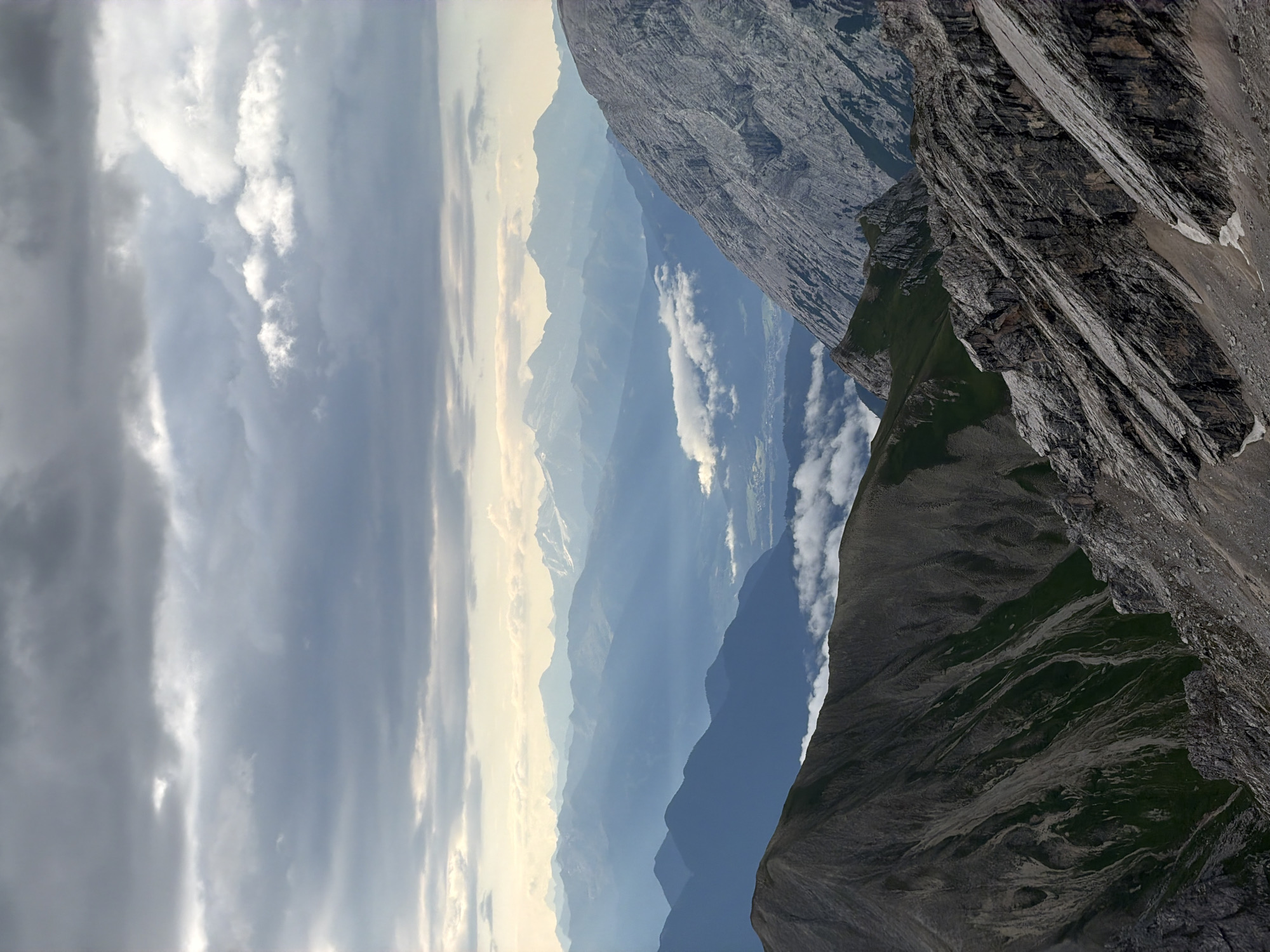 The image displays a panoramic view of a mountain range under a cloudy sky. In the foreground, rugged, rocky slopes dominate the scene, with some patches of green vegetation. The middle ground features a valley with several layers of receding mountains, fading into a hazy blue hue. The background reveals distant, snow-capped peaks. A few clouds are visible in the valley, adding depth to the landscape. The overall impression is one of vastness and natural beauty. The location is identified as the Zugspitze mountain, the highest peak in Germany, near Garmisch-Partenkirchen. No people or specific objects are visible in the image, only natural elements.