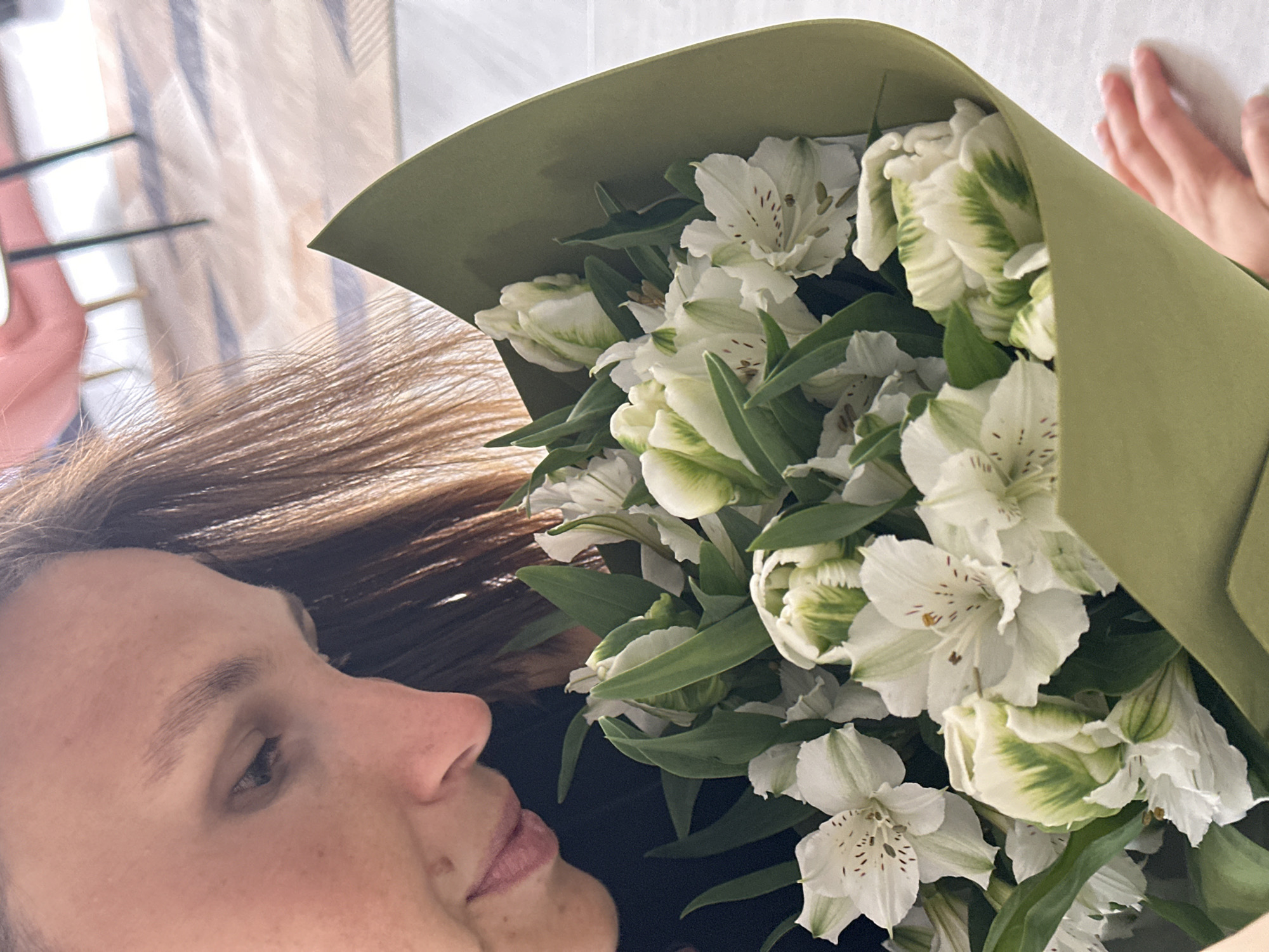 A person is holding a bouquet of white alstroemeria flowers wrapped in green paper. The person has brown hair and fair skin, and their face is in profile. The person's left hand is visible behind the bouquet. The background is a blurry interior, possibly a home. The location is not evident in the image.
