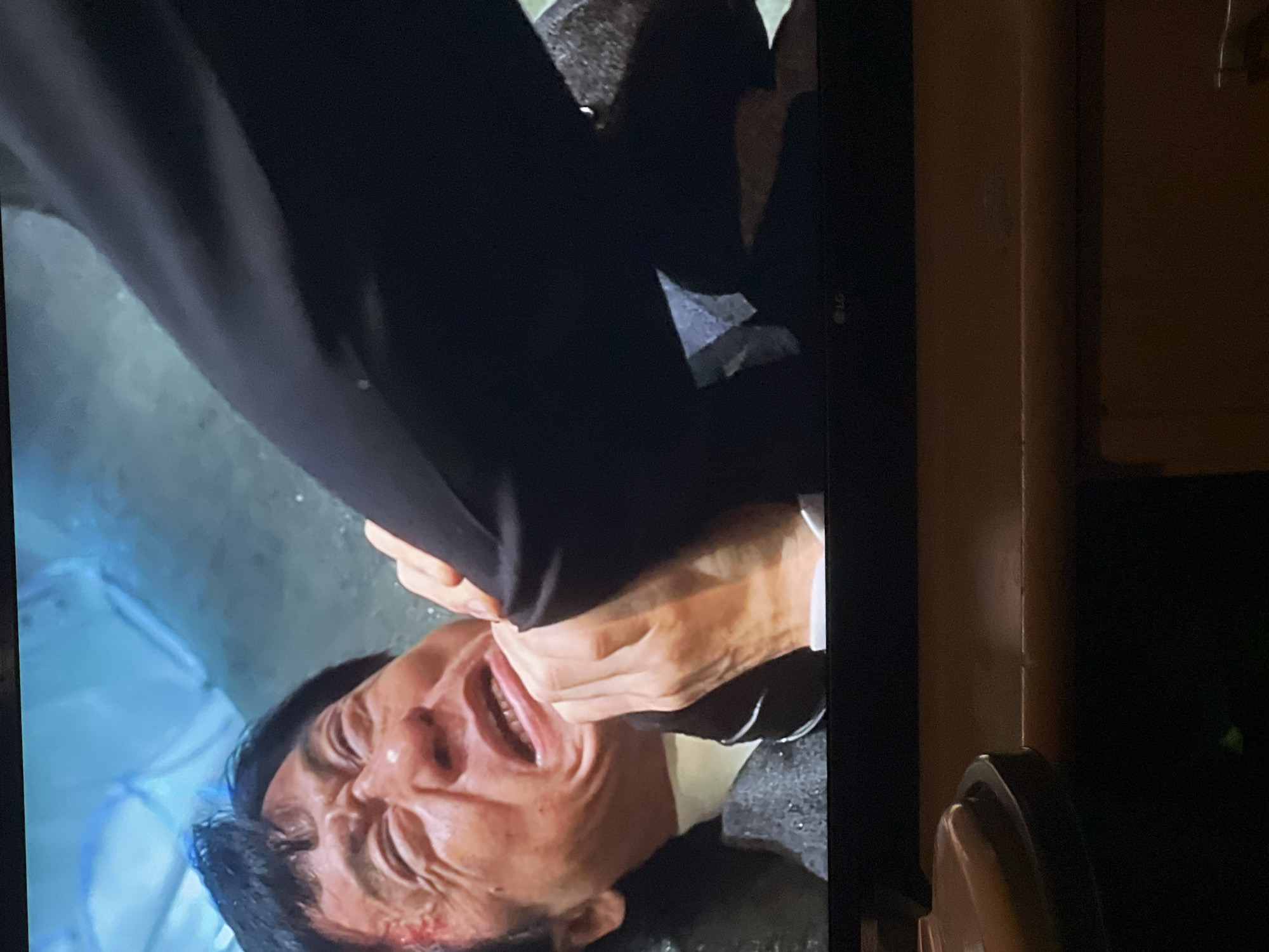 The image displays a close-up of an East Asian male, identified as actor Jackie Chan, caught in a physical struggle. His face is visibly contorted with intense pain or exertion, mouth agape, revealing teeth. A distinct red abrasion or laceration is present on his left temple. He is dressed in a dark, textured suit jacket over a light-colored collared shirt. His right hand is firmly gripping the dark trousers of an unseen individual's leg, which is angled across the foreground, appearing to restrain or apply pressure to him. The background consists of dark, wet, or reflective concrete surfaces, indicating a utilitarian, possibly subterranean, or industrial environment. A crumpled light blue object, resembling plastic sheeting or a tarp, is visible in the upper left portion of the background. The scene depicts a violent interaction occurring in a dimly lit, enclosed space. This generic industrial interior is located within the city of Sofia, Bulgaria.