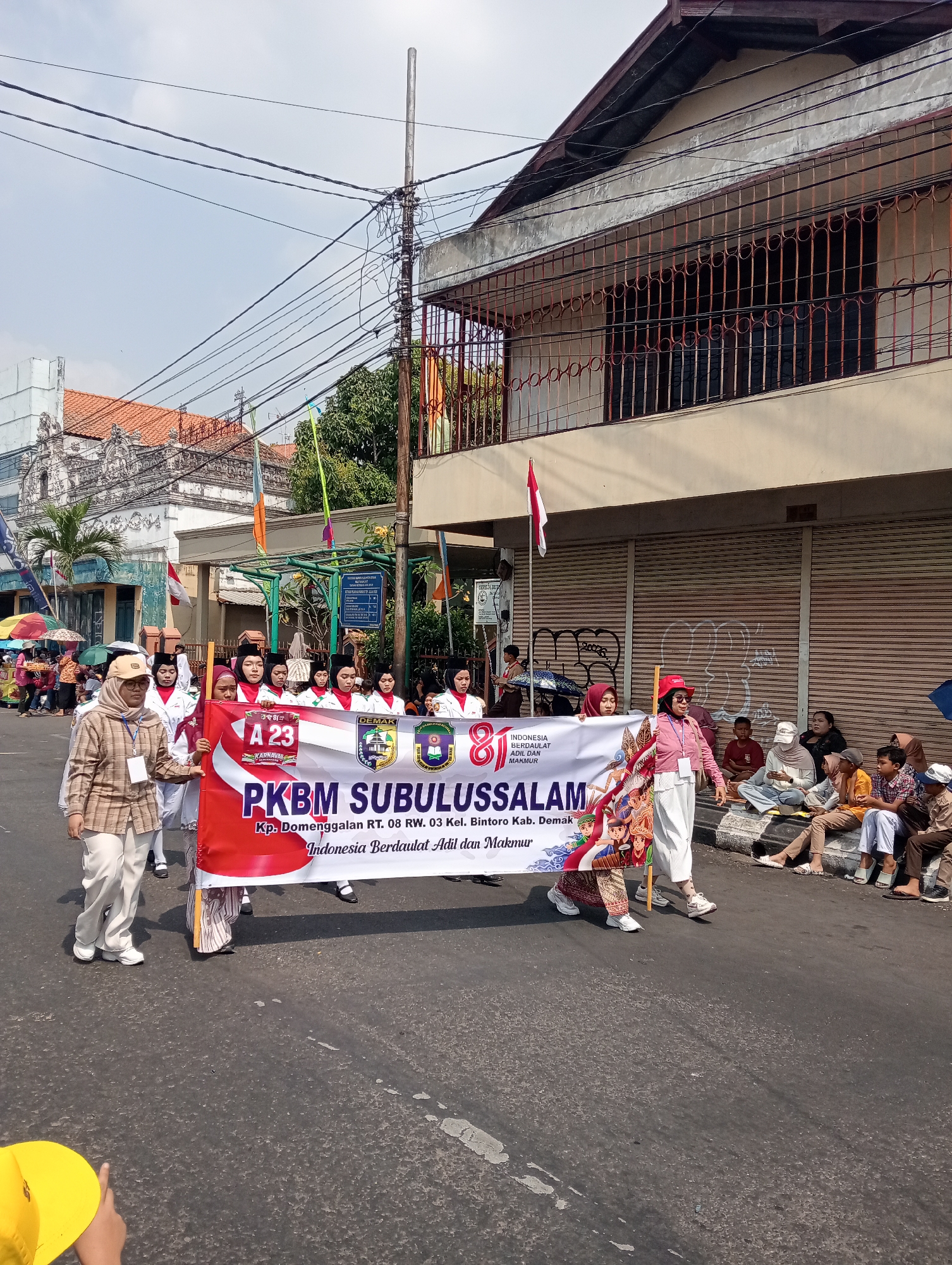 A group of people, predominantly young women in uniform, are participating in a parade. They are holding a banner that reads "PKBM SUBULUSSALAM" and displays "INDONESIA BERDAULAT ADIL DAN MAKMUR". The banner also includes the address "Kp. Domenggalan RT. 08 RW. 03 Kel. Bintoro Kab. Demak". Some individuals in the foreground are walking and holding the banner, while others are seated on the curb watching the parade. The parade is taking place on a street lined with buildings, some of which have awnings and decorative metalwork. Overhead, electrical wires are strung across the street. The sky is visible and appears to be partly cloudy. The overall scene depicts a community event or celebration in Bintoro, Indonesia.