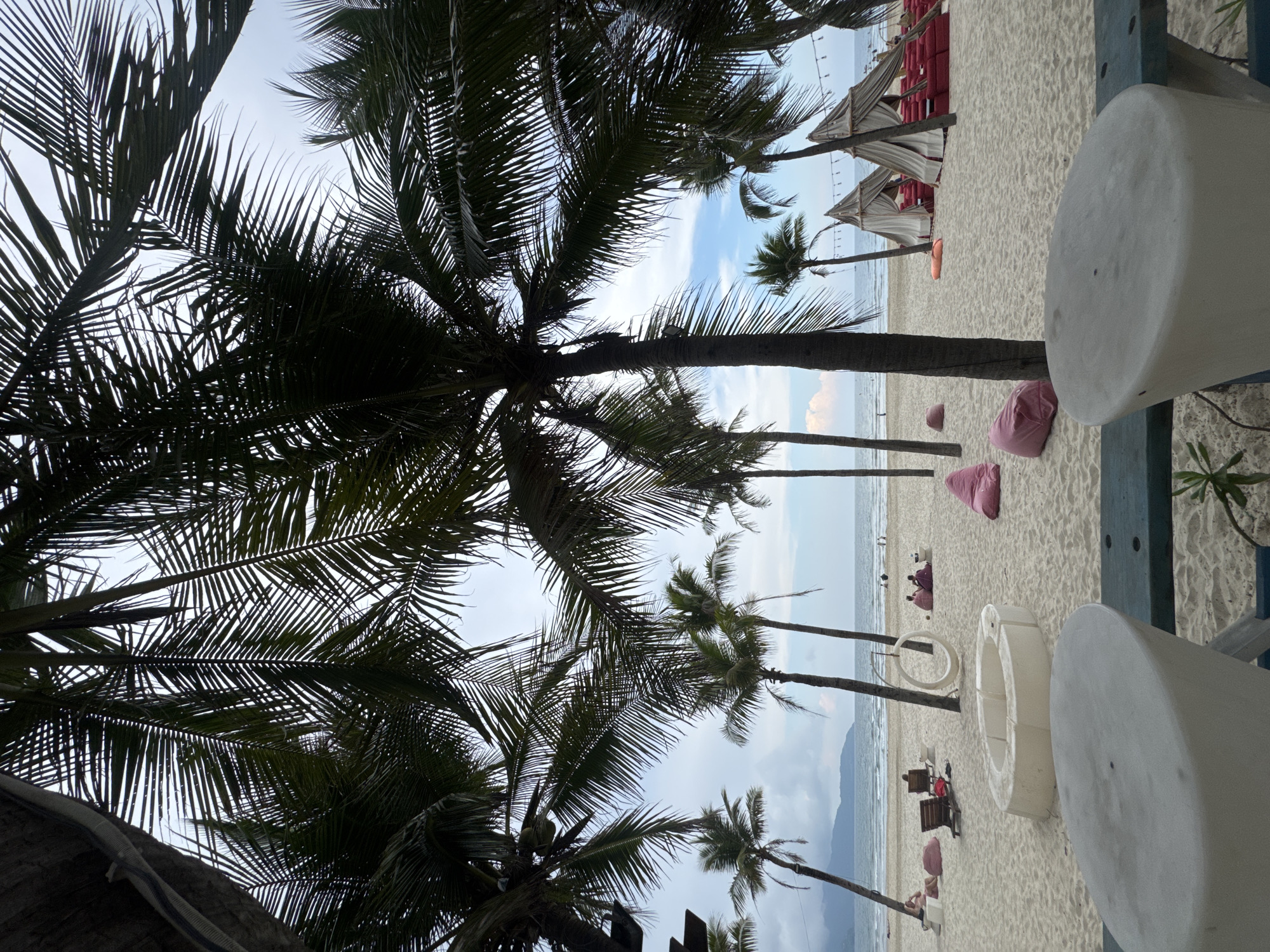 The image shows a beach scene in Đà Nẵng, Vietnam, dominated by palm trees framing the sky. The sand is light-colored and sparsely populated with people relaxing in beach chairs and on beanbag cushions. A line of covered lounge chairs stretches along the shoreline. Two large, white, cylindrical objects are visible in the foreground, possibly part of a beachfront bar or dining area. The sea is calm and the sky overcast, suggesting a cloudy day.