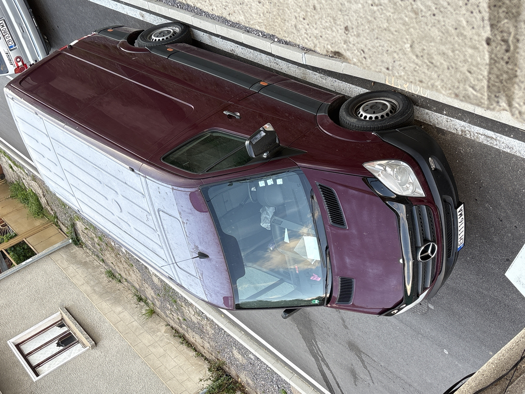 An overhead, slightly angled, medium shot captures a parked, dark red Mercedes-Benz Sprinter van on a paved street in Anton, Bulgaria. The van is positioned parallel to a raised concrete curb with double white painted lines indicating a parking area. To the left of the van, a building with a light-colored, textured facade and a window with white curtains is visible. A small amount of overgrown vegetation lines the base of the building and extends towards the street. The van's windshield reflects a pale light, and a person's blonde hair is visible through the glass. The license plate of the van reads "EH1116MA". Behind the Sprinter, a silver car is partially visible, with a license plate that appears to read "CB 0380 MX". The overall environment suggests a residential or commercial street setting.