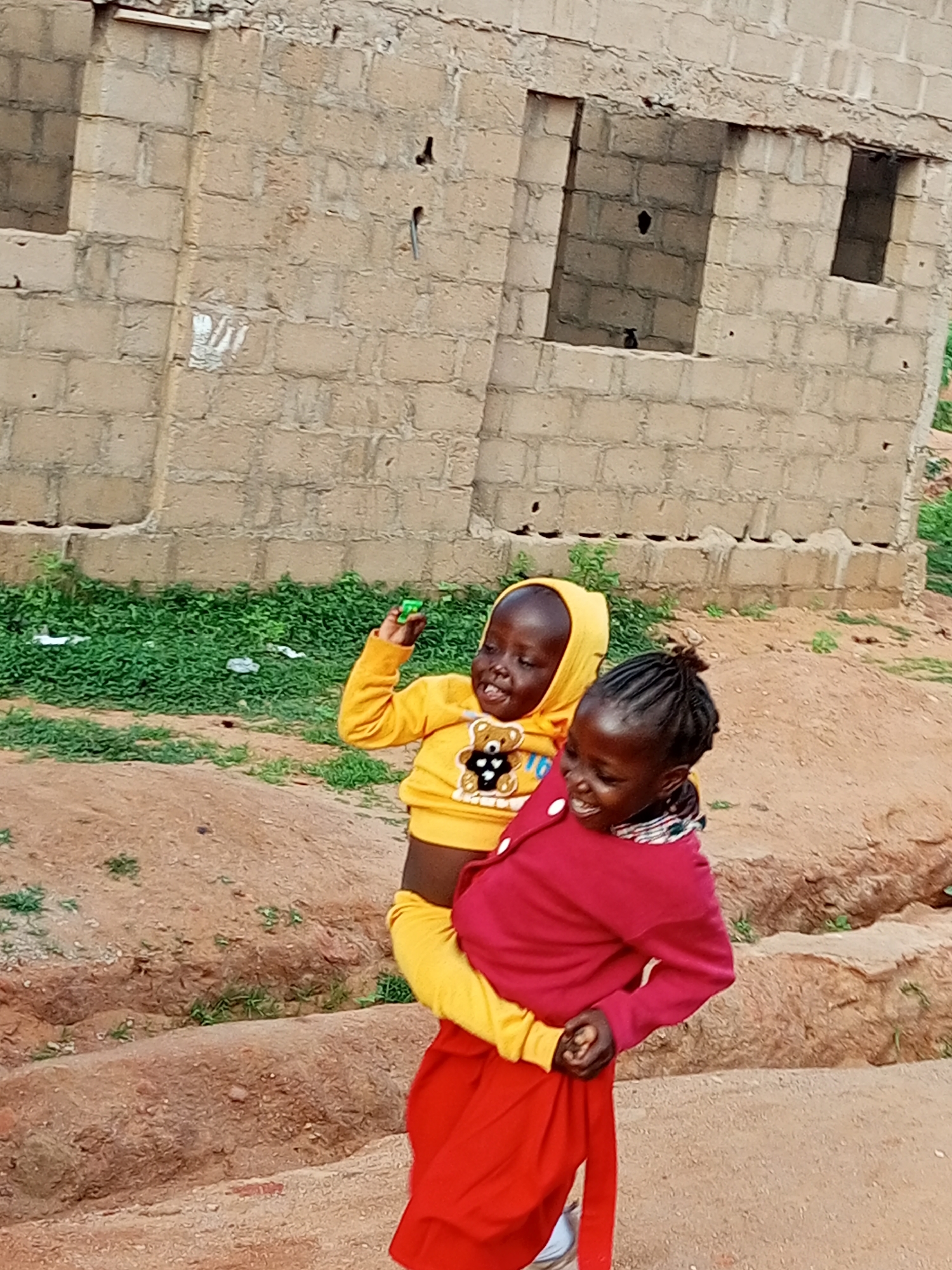 In Tumfure, Nigeria, two young children are captured in a moment of shared joy. The child in the foreground, wearing a red sweater and a flowing red skirt, carries a younger child on their hip. The younger child, clad in a bright yellow hoodie with a teddy bear graphic, beams with a wide smile and holds a small green object in their raised hand. They appear to be standing on a dirt path in front of a partially constructed building made of light-colored bricks. The background features a dense growth of green vegetation and the rough texture of the unfinished concrete structure. The scene is outdoors, under natural light.