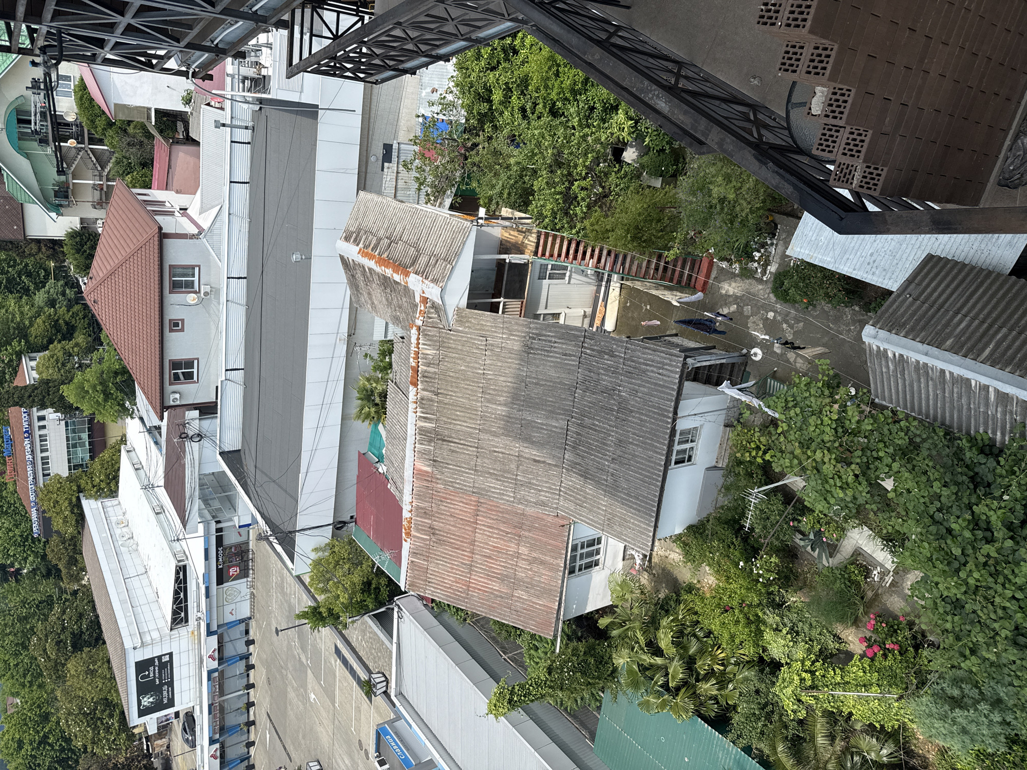 An aerial view shows a dense urban landscape in Sochi, Russia, characterized by a mix of residential buildings, commercial structures, and abundant greenery. In the foreground, a weathered, multi-story residential building with a corrugated metal roof and a red-tiled roof section dominates the scene. An external staircase with red steps leads up to a balcony. Adjacent to this structure is another building with a similar gray, corrugated roof.
Behind these residential buildings, a large, modern commercial complex with a white facade and large windows is visible. Its roof is flat and dark, with visible air conditioning units. Further in the background, more buildings of various architectural styles, including a prominent white building with a sign indicating "РЕКЛАМА ФОТОГРАФИЯ" (Advertising Photography), are scattered amongst lush green trees. The image captures a typical view of Sochi, a coastal city known for its subtropical climate and extensive vegetation intertwined with urban development. The overall impression is one of a lively, densely populated area with a significant presence of trees and foliage.