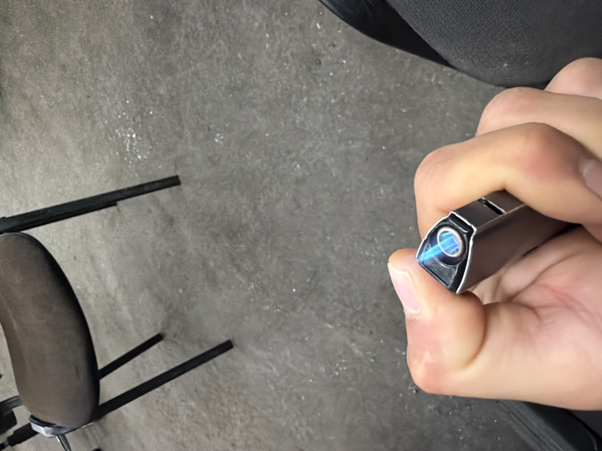A close-up shot shows a person's hand holding a silver electric lighter with a blue flame at the tip. The background consists of a rough gray surface, possibly concrete or asphalt, with small white specks. To the upper left, a portion of a dark brown chair with a worn seat and black metal legs is visible, suggesting an indoor or outdoor seating area. The scene does not contain any identifiable landmarks or elements that would indicate its specific location in Burgas, Bulgaria.