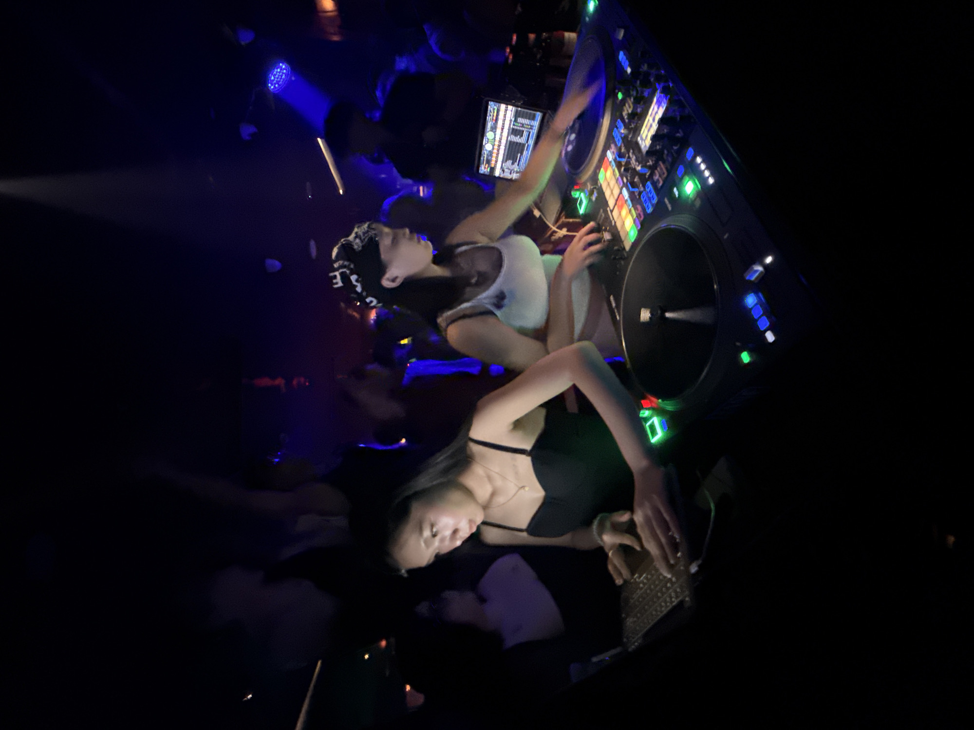 Two female DJs are performing at a nightclub in Liangjiang, China. The DJ on the left is wearing a black dress and operating a laptop, while the DJ on the right is wearing a white tank top and a bandana, and is using a DJ console with turntables and a mixer. The nightclub is dimly lit with blue and purple lights, and there are people in the background, some of whom are dancing. The overall atmosphere is energetic and festive.