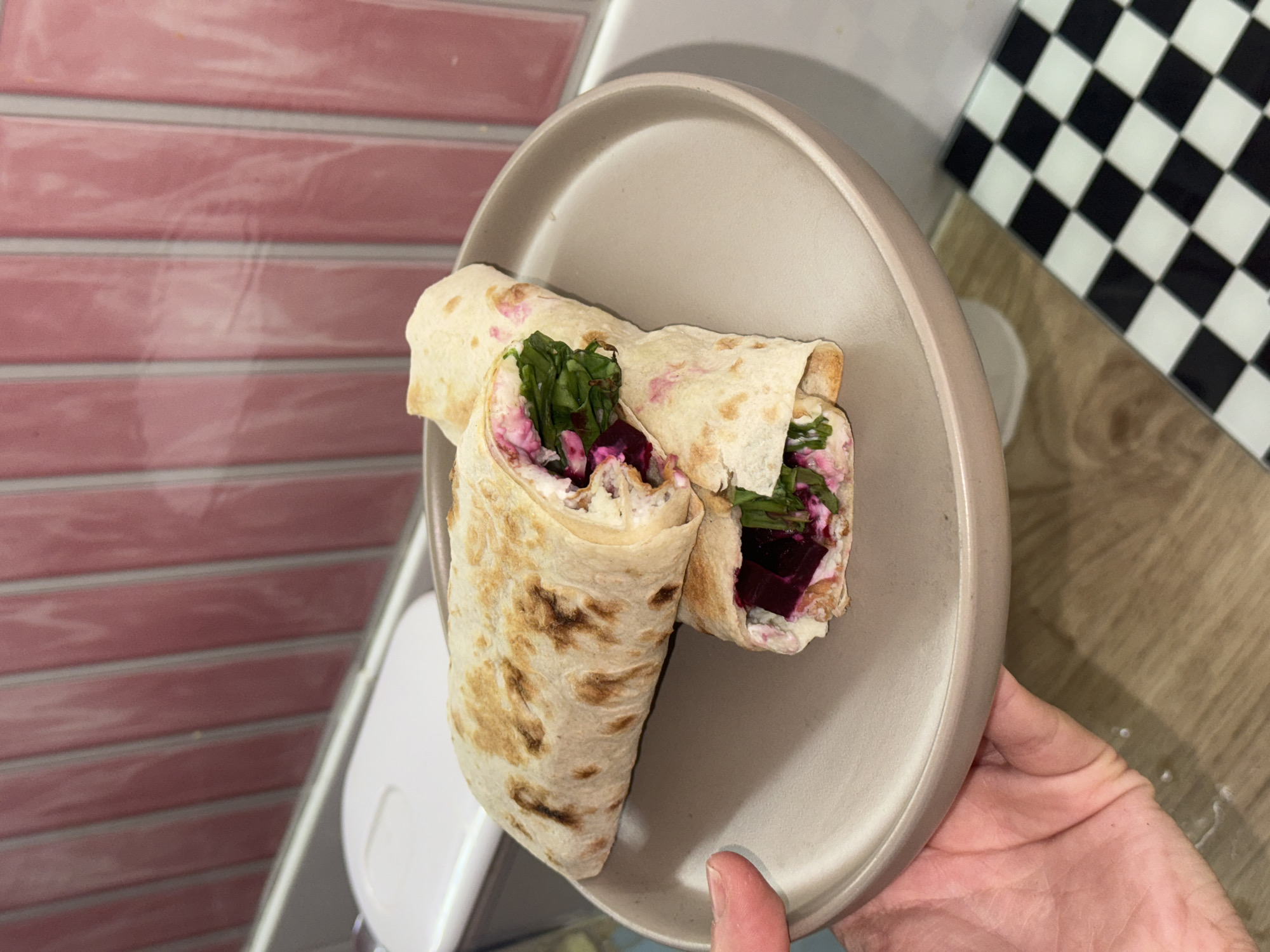 A person holds a light-gray plate with two wraps on it. The wraps appear to be made of flatbread and contain a filling that includes greens, beets, and a white sauce. The hand holding the plate is Caucasian, and the thumb is visible. The background includes a pink-tiled wall on the left and a black and white checkered surface on the top right, likely a countertop or table. Below the plate, there is a light-colored wooden surface. The location cannot be determined to be Krasnoyarsk, Russia from the image.