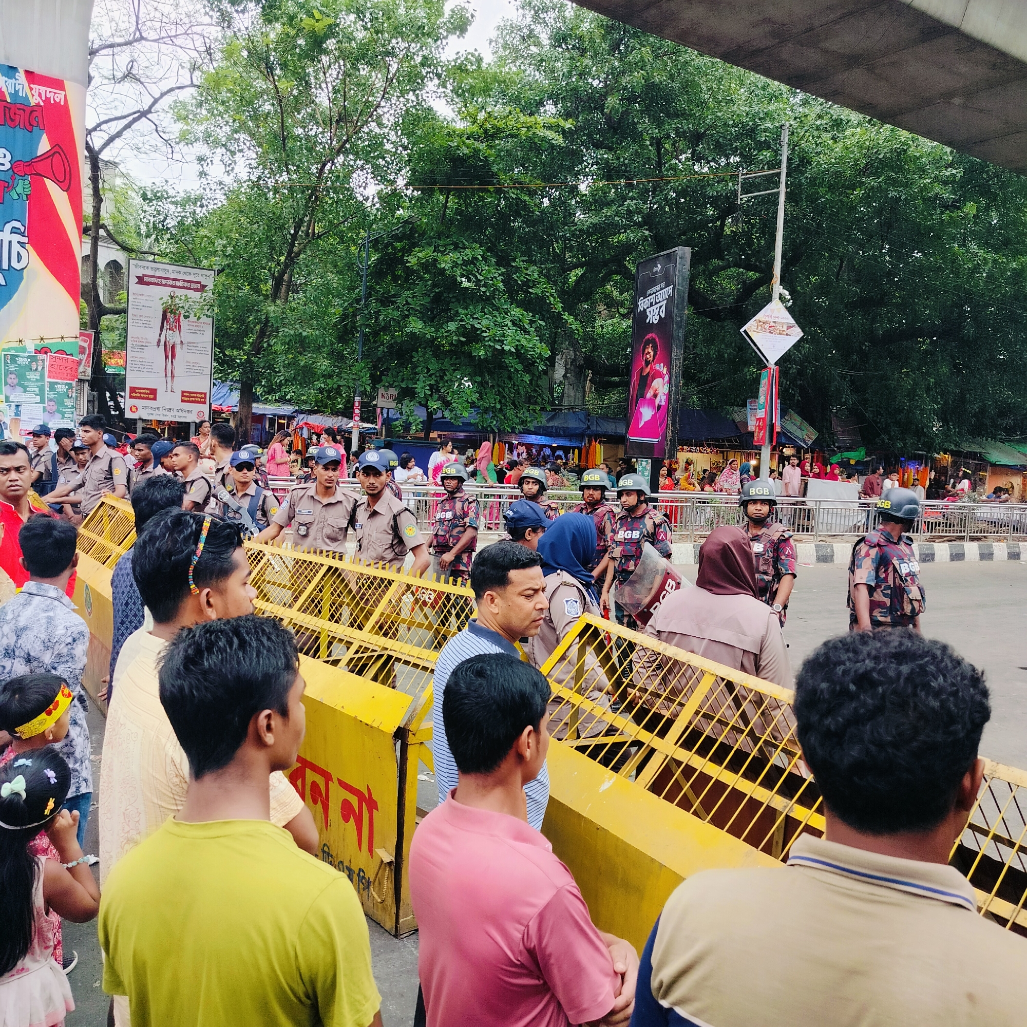 The image shows a crowd of people in Dhaka, Bangladesh, standing behind a yellow barricade. Behind the barricade are security personnel, some wearing helmets and camouflage uniforms marked with "BGB" (Border Guard Bangladesh). The people in the crowd are dressed in various casual clothes. Overhead, there is a structure, possibly a bridge or elevated roadway.
In the background, there are trees, banners, and posters with text in Bengali. The posters include images of people and informational graphics, such as a human anatomy illustration. There are also commercial advertisements.