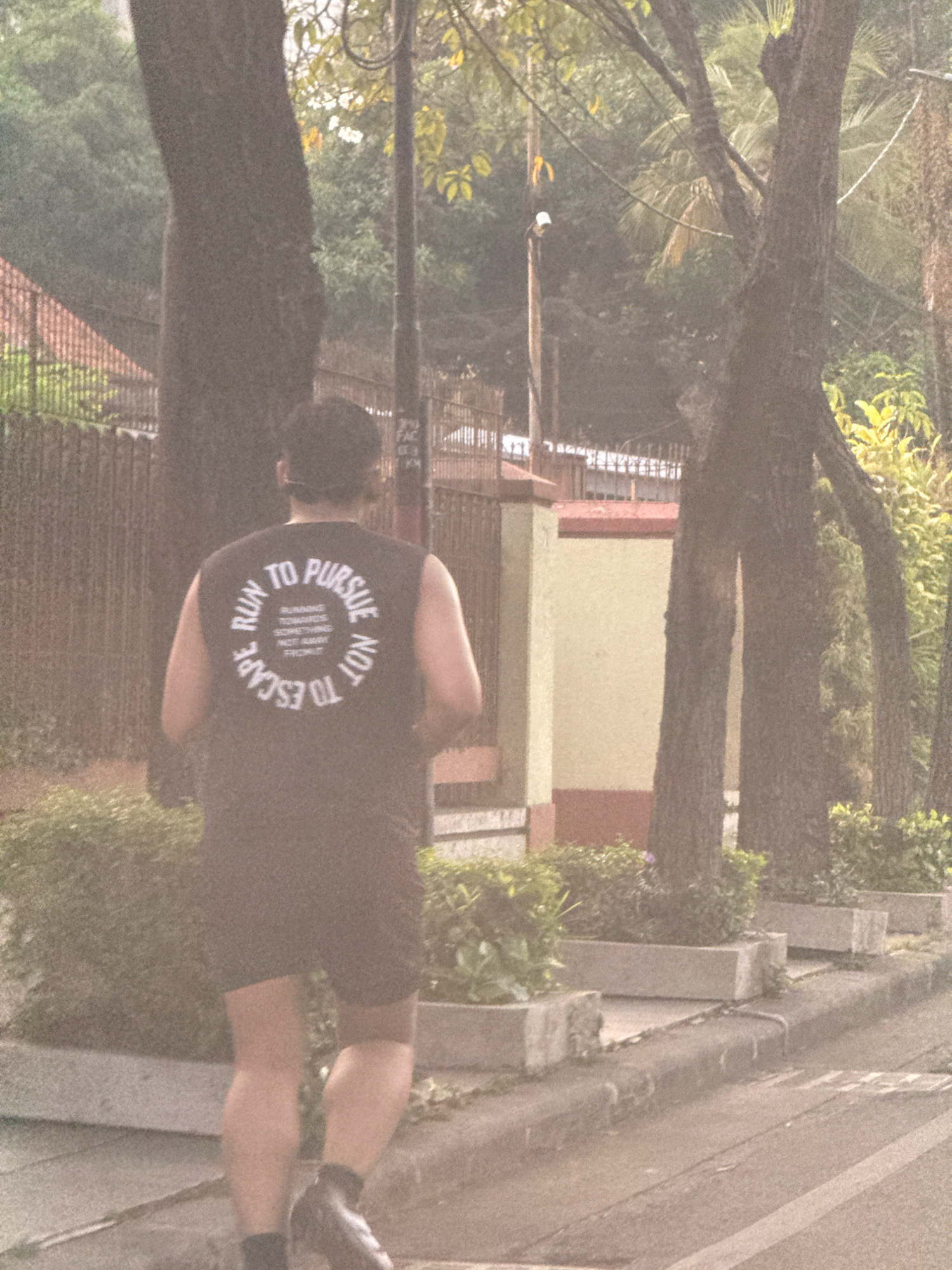 A person, facing away from the camera, is jogging along a sidewalk in Surabaya, Indonesia. They are wearing a black sleeveless shirt with white text that reads "RUN TO PURSUE NOT TO ESCAPE" in a circular pattern, with smaller text inside the circle. The person is also wearing black shorts and dark shoes with black socks. The environment appears to be a street lined with trees and planters. In the background, there is a building with a light-colored facade and red trim, and a fence. The overall lighting suggests it is daytime, with diffused light, possibly indicating an overcast sky or early morning/late afternoon.