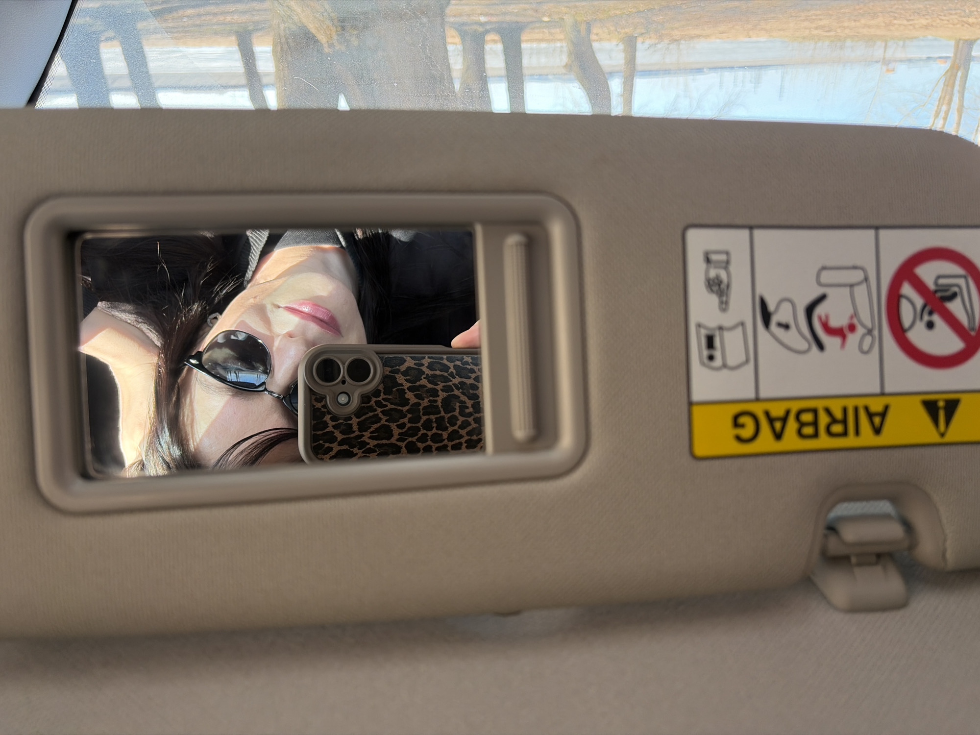 An upward-facing, eye-level shot captures a person taking a selfie inside a car in Chaikovsky, Russia. The central focus is a woman's reflection in the vanity mirror of a beige car sun visor. She has long dark hair, light skin, and is wearing sunglasses pushed up onto her head, reflecting the bright outdoor light. Her lips are painted a glossy pink, and she has a subtle smile. She holds a smartphone with a distinctive leopard-print case, which is also reflected in the mirror, obscuring part of her chin.
To the right of the mirror, on the sun visor, there is an upside-down yellow warning label with black text that reads "AIRBAG." Below this text, several pictorial warnings are visible, including symbols related to airbag deployment, the use of a child seat, and an instruction to consult a manual.
Above the sun visor, through the car's windshield, the reflection of a sunny, clear daytime environment is visible. Bare trees and what appears to be a body of water or distant landscape are reflected, suggesting late autumn, winter, or early spring. The overall impression is a casual, sunlit moment captured within the vehicle.