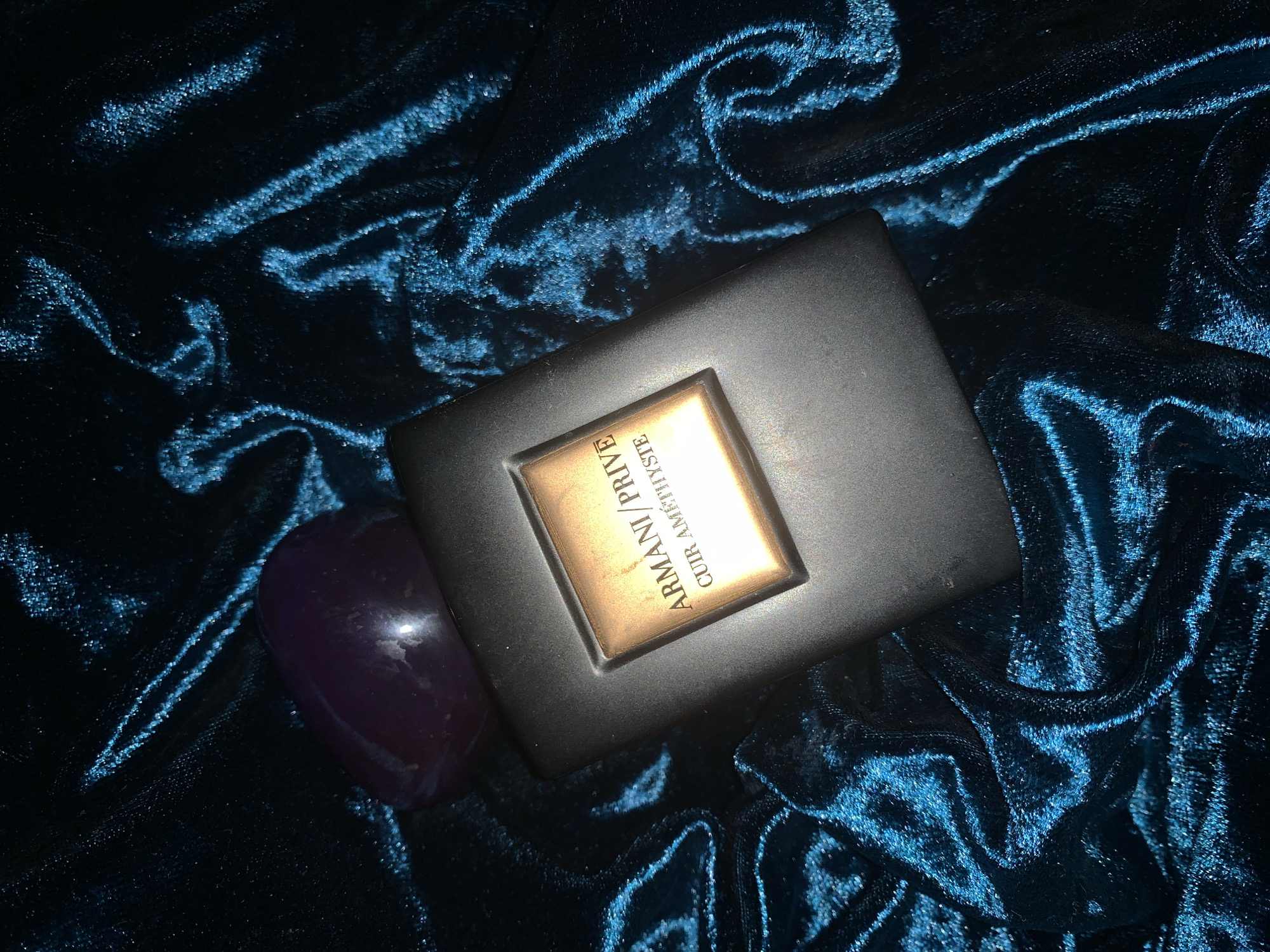 The scene depicts a rectangular black box labeled "ARMANI/PRIVE CUIR AMETHYSTE" resting on a crumpled, deep blue velvet fabric. A polished, purple stone is situated next to the box. The location and city are unknown.