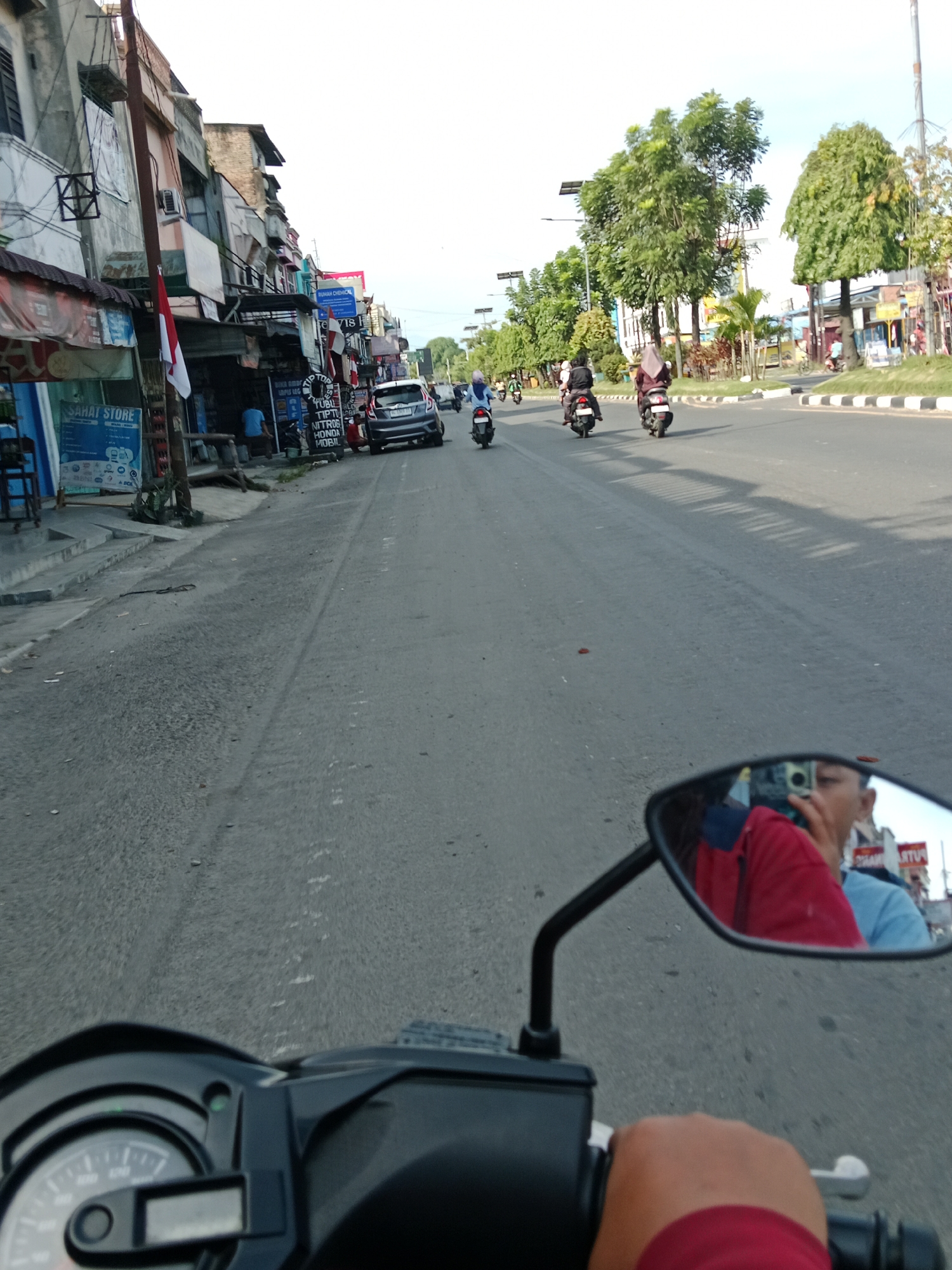 The image captures a street scene in the city of Binjai, Indonesia, likely during the daytime. The perspective is from a motorcycle, showing the handlebars and a portion of the instrument panel in the foreground. A side mirror reflects the rider, who is wearing a red shirt, and another person wearing a blue shirt. The street is paved and appears to be a two-lane road with traffic. Several motorcycles are visible on the road, some with riders. On the left side of the street, there are buildings with shops, including one with a sign that says "SAHAT STORE." A red and white Indonesian flag is hanging from a pole. On the right side of the street, there are trees and more buildings in the distance. The overall atmosphere suggests a busy urban environment.