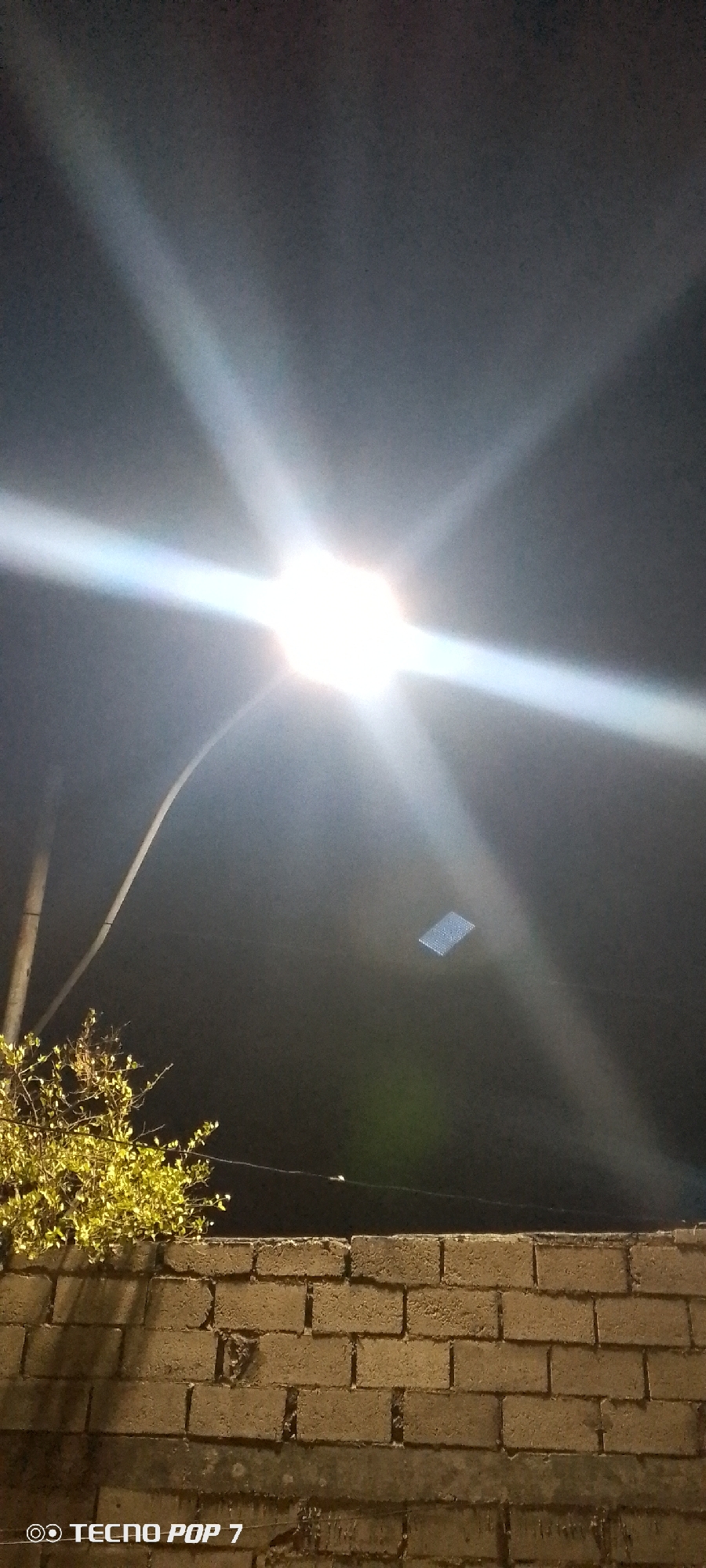 An image of a nighttime scene, likely in Altagracia, Venezuela, features a bright streetlight shining prominently against the dark sky. The light flares outwards, creating a starburst effect. Below the light, there's a glimpse of what seems to be a rectangular panel or object, perhaps a solar panel, suspended in the air. A curved pole, presumably holding the streetlight, is visible to the left. A leafy bush or small tree sits on the left side of the frame, partially obscuring a brick wall that spans across the lower portion of the image. The wall is made of rough-textured bricks. A thin wire stretches horizontally across the upper part of the frame, behind the wall. The "Tecno Pop 7" watermark is present in the bottom left corner of the image.