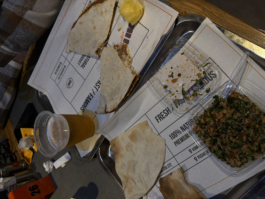 Two metal trays hold various food items. The first tray contains pieces of flatbread, a slice of lemon, and a drink in a clear plastic cup. The second tray features two clear plastic containers holding salad. The food is placed on white paper with black and orange writing, including "FRESH / FAMOUS," "100% NATURAL PRODUCT," and a phone number. A portion of a garment, possibly plaid, is visible in the upper left. A small orange tag with "26" printed in white is at the bottom left.