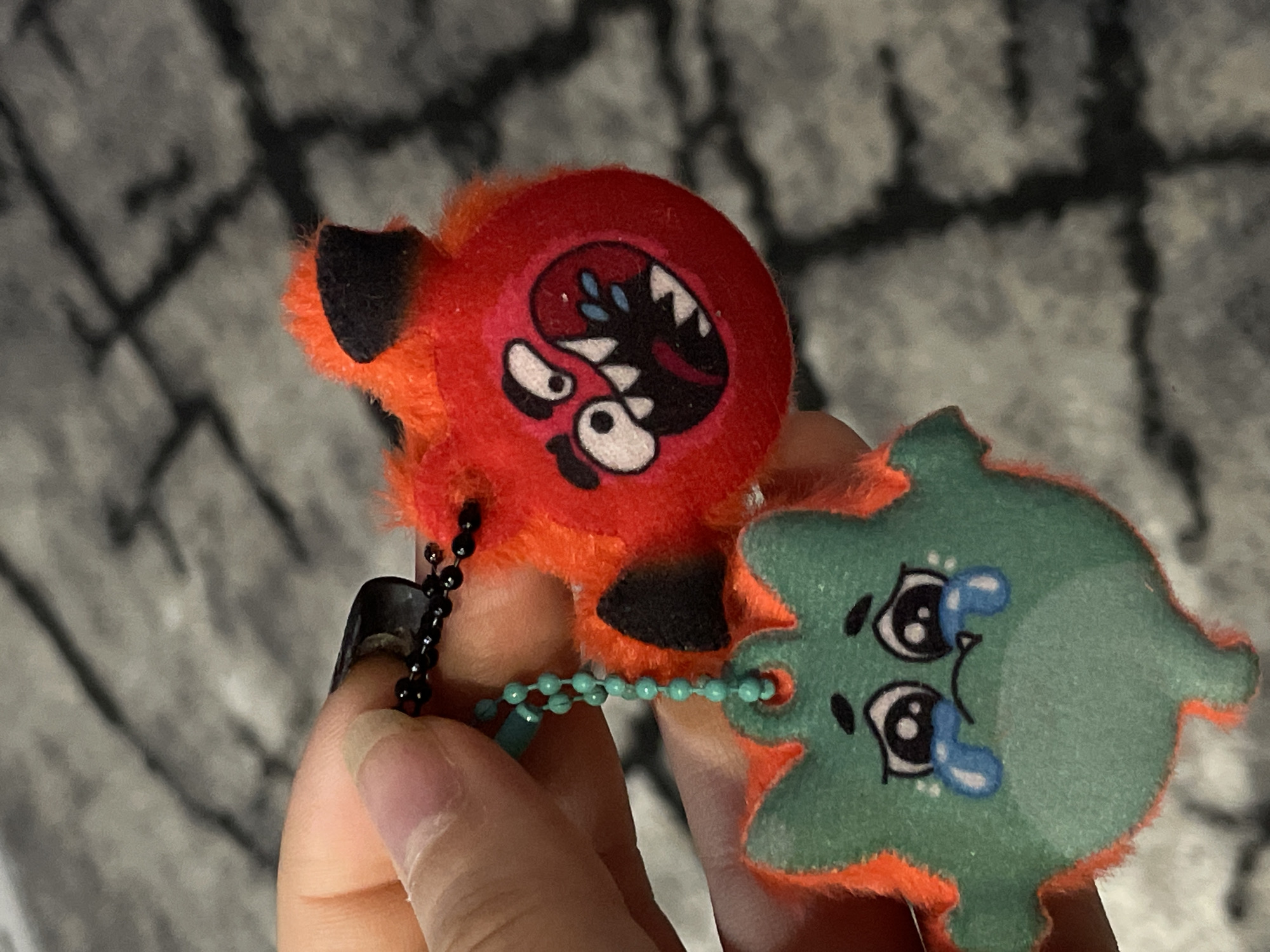 A hand holds two plush keychains. The keychain on the left is predominantly red and orange with a cartoonish face featuring a gaping mouth with jagged teeth and an angry expression. This keychain is attached to a black ball chain. The keychain on the right is green and orange with a sad face, complete with drawn tears. These two keychains are connected by a turquoise ball chain. The background is a blurred, grayish-black pattern. There is no indication of the city Chaikovsky, Russia.