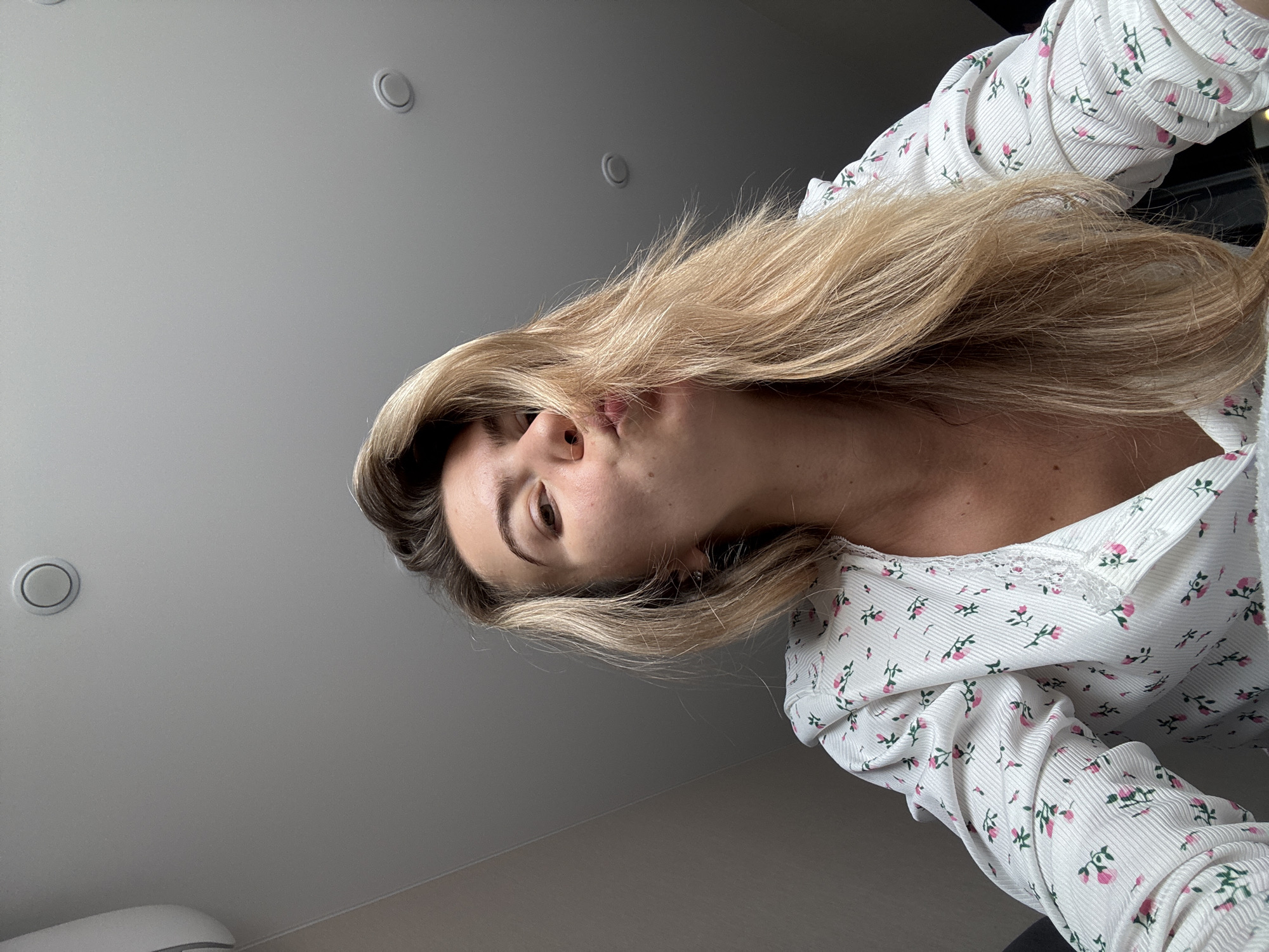 A woman is visible in the image, wearing a white top with a pink floral pattern and lace trim. Her long, blonde hair is styled with some strands falling across her face. She appears to be looking directly at the camera with a slightly pursed expression. The background is a gray ceiling with three round, recessed lights. The location is Unknown, Unknown.