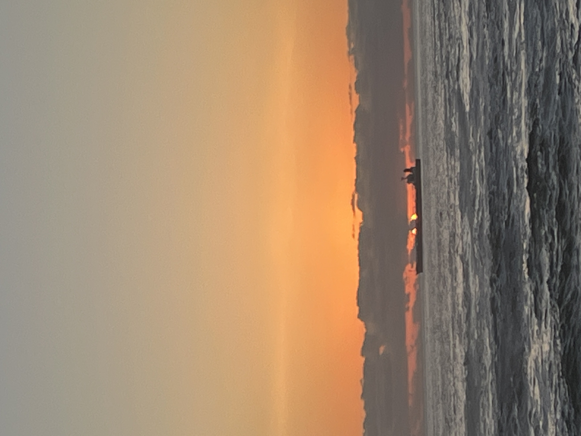 The image shows a seascape at sunset, likely near Arica, Chile given the request. The right side of the image contains water with visible wave patterns. Towards the center, the sun appears on the horizon, partially obscured by clouds or haze. Silhouetted figures are faintly visible on the water near the sunset. The left side features a diffuse, gray-orange sky gradient.