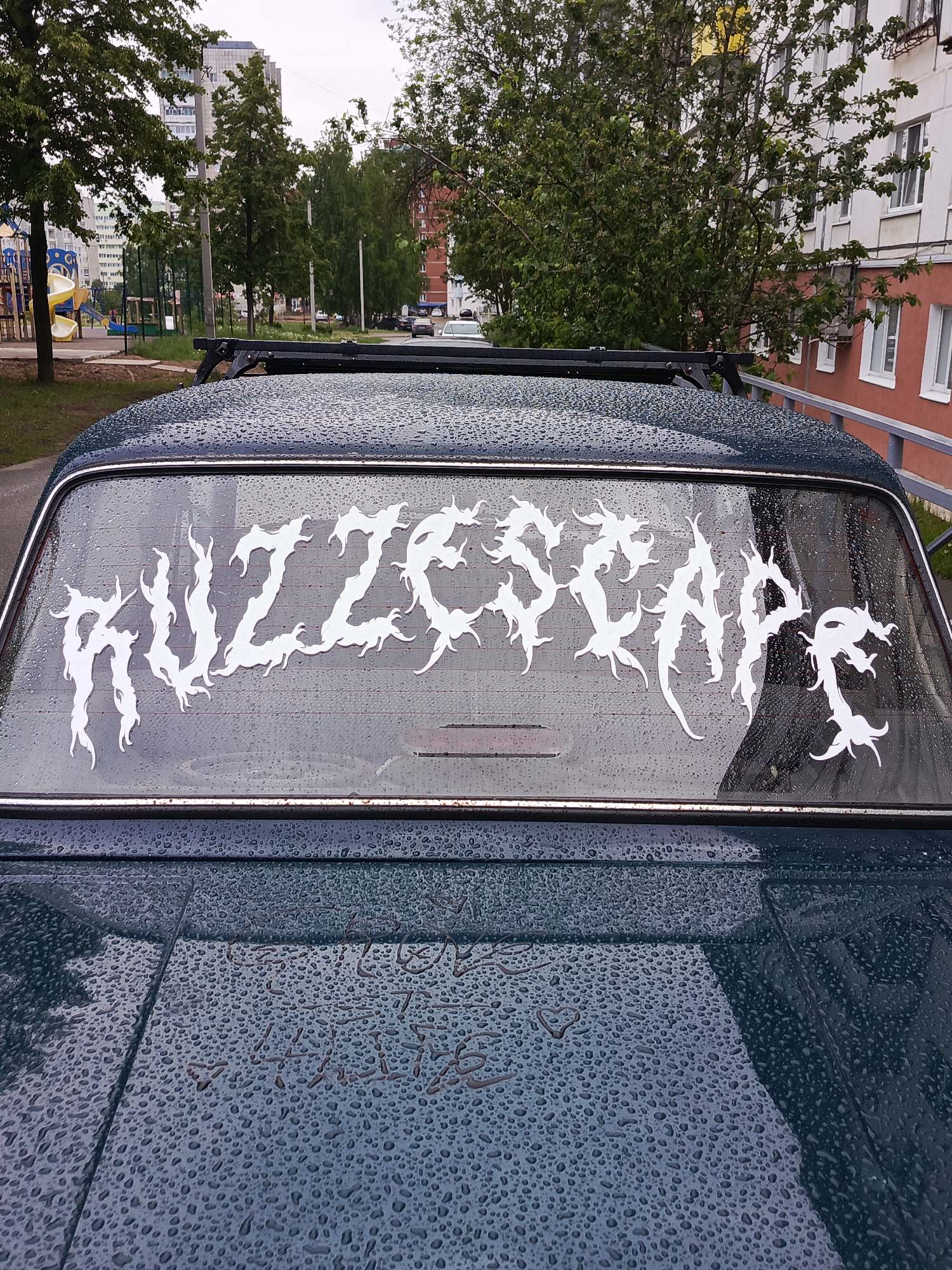 A close-up shot depicts the rear window of a dark-colored vehicle, likely a sedan, parked on a street. The window is covered in water droplets, indicating recent rain or wet conditions. Affixed to the inside of the rear window is a large, white decal that reads "RUZZESCAPE" in a stylized, aggressive font with jagged edges, resembling metal band logos.
The roof of the car, visible above the window, has a roof rack installed. More water droplets cover the car's surface.
The background reveals an urban environment. There is a playground with colorful equipment visible to the left, and a multistory apartment building in the distance, along with trees and other greenery.
The scene is located in the city of Chaikovsky, Russia.