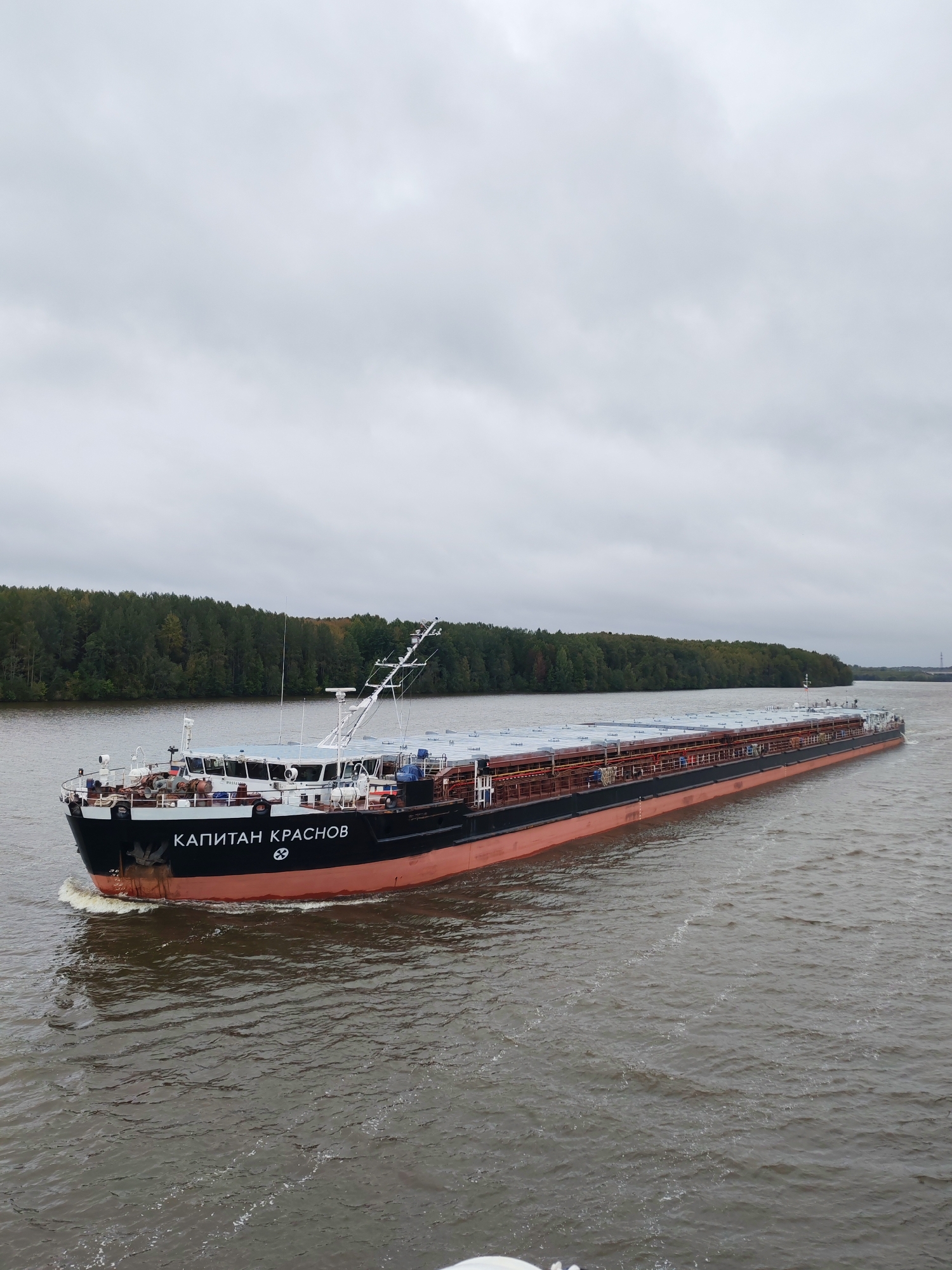 A large, long cargo barge named "КАПИТАН КРАСНОВ" (Kapitan Krasnov) travels down a wide, murky river under an overcast sky. The barge is black with a distinctive orange-red hull and is loaded with rows of silver-covered cargo containers. A white superstructure with windows is visible at the front of the barge, housing the navigation bridge. A tall mast with antennae extends upwards from the superstructure. The barge is creating a foamy wake as it moves. The riverbanks are lined with a dense forest of green trees, with some hints of autumnal colors. The overall scene suggests a calm, possibly cool, day on a navigable waterway. The location is identified as Kosye Gryady, Russia.