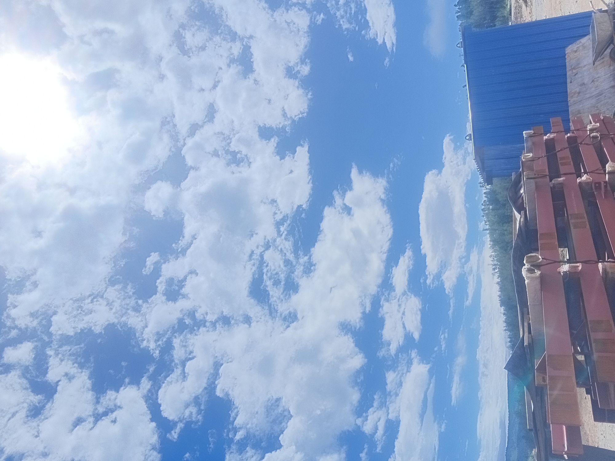 The image displays an outdoor scene under a bright, partially clouded blue sky. In the foreground, a stack of large, reddish-brown metal beams is visible. To the right of the beams, a corrugated blue metal shed or structure stands. The background features a dense forest on a sloping terrain, with a hazy mountain range in the distance. The ground appears to be rough and unpaved, suggesting a construction or industrial area. A wheelbarrow is partially visible in the bottom right corner. The overall impression is one of a partly developed or industrial site under a sunny sky. There are no people or animals present in the image. The location is identified as Srednyaya Kiya, Russia.