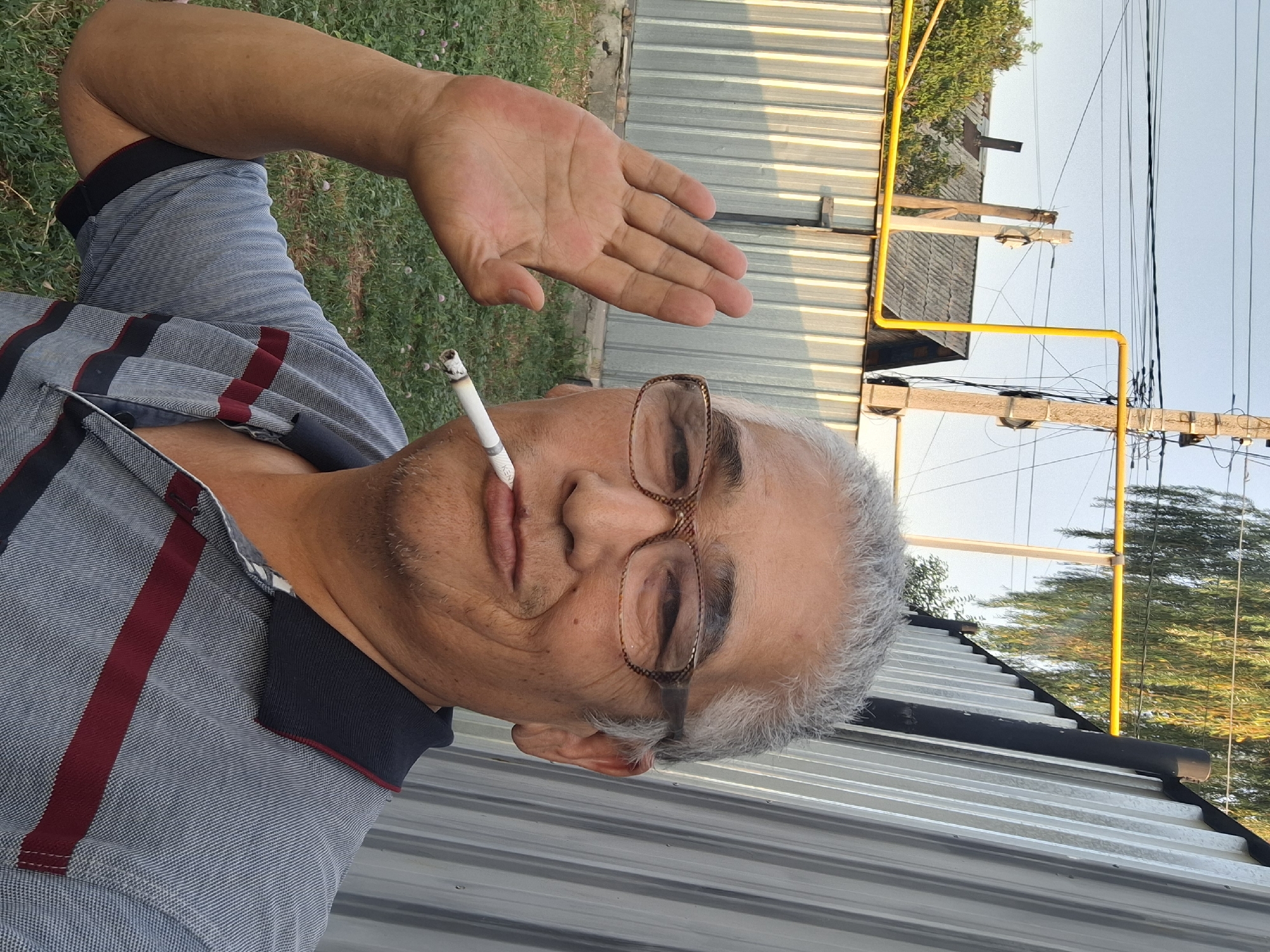 An eye-level, medium shot shows a middle-aged East Asian man holding his left hand up in a greeting or stop gesture, his palm facing the camera. He is wearing glasses with patterned frames and a grey polo shirt with navy and burgundy stripes. He is smoking a cigarette, which is held between his lips. The man's hair is grey and receding. The background consists of a corrugated metal fence, grass, and some trees and power lines, suggesting an outdoor, possibly rural or semi-urban setting. The lighting suggests it is late afternoon or early evening. The presence of a yellow gas pipe running along the fence and power lines indicates that this is a residential area. Based on the visual cues, such as the architecture of the houses visible in the background and the general aesthetic, this scene is likely located in the city of Almaty, Kazakhstan.