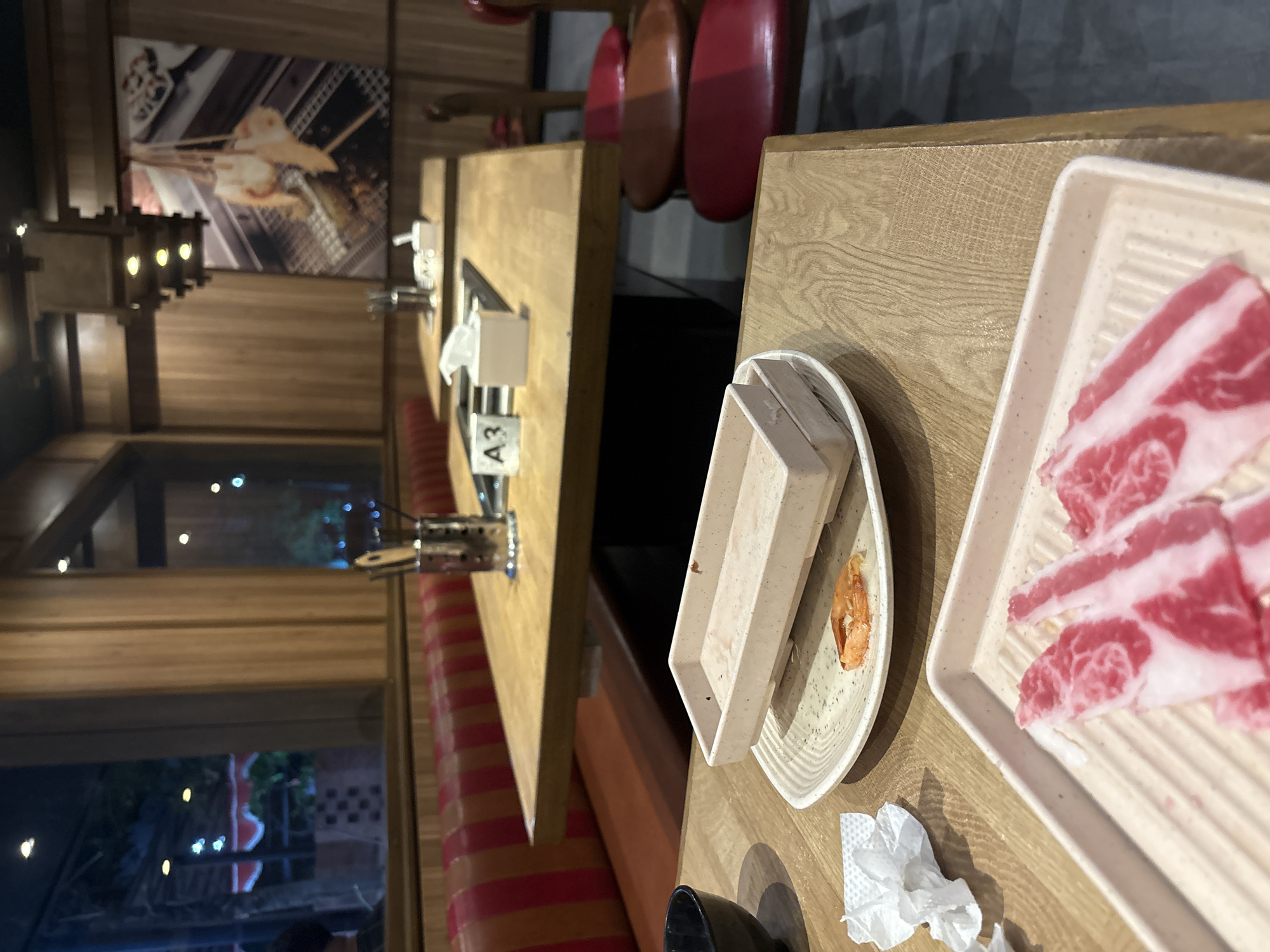 The image depicts a table setting at what appears to be a restaurant in Cirebon, Indonesia. The table is made of wood and is set with various dishes. In the foreground, there is a tray with raw, thinly sliced red meat, likely beef, with visible marbling. Beside it, a white paper napkin is crumpled. Further back on the table, a plate holds a single cooked shrimp, and a rectangular white tray with some residue is stacked on top of it. Another table is visible in the background, with a similar setup and a sign labeled "A3". The restaurant interior features wooden paneling on the walls and booths with red and orange striped upholstery. A decorative light fixture hangs from the ceiling. Through a window in the background, a glimpse of an outdoor area with foliage and lights can be seen. The scene suggests a meal is in progress or has recently concluded.