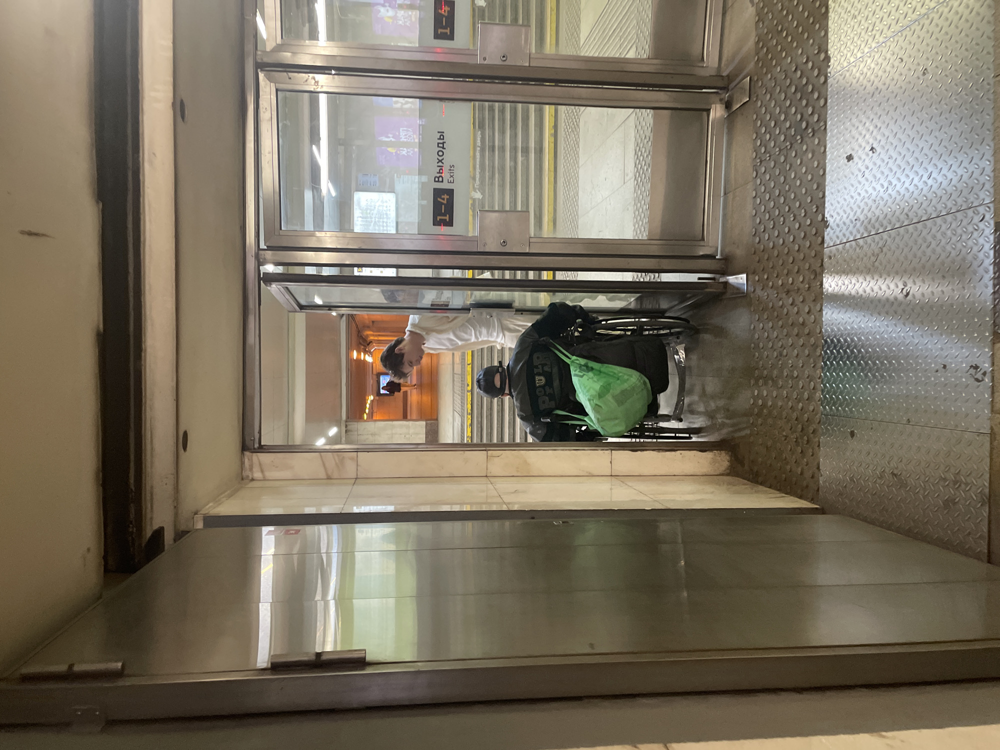 A person in a wheelchair is being assisted by another person through a set of automatic glass doors at what appears to be a subway station. The person in the wheelchair is wearing a black jacket and a black cap, and has a bright green bag on their lap. The other person is wearing a white shirt. Behind them, stairs can be seen leading down, and signage in Russian and English indicates "Выходы" (Exits) and "1-4". The scene is indoors, with tiled walls and metal flooring visible. The lighting suggests it is daytime. The image is oriented vertically.