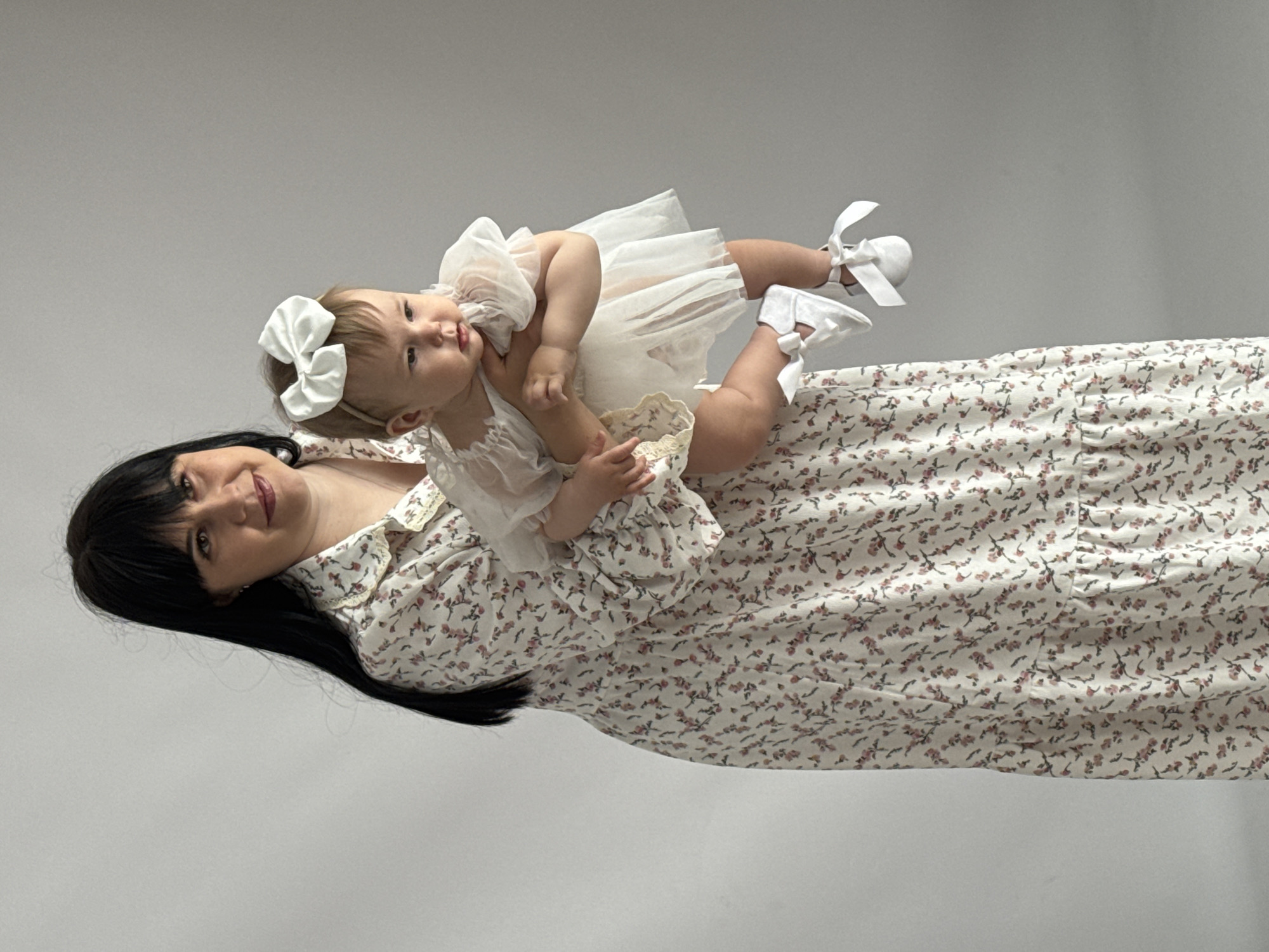 A woman with dark hair holds a baby in a white dress with matching shoes and a bow. The woman wears a floral-patterned dress. The background is a solid, light gray color. There is no evidence to suggest this image was taken in Izhevsk, Russia.