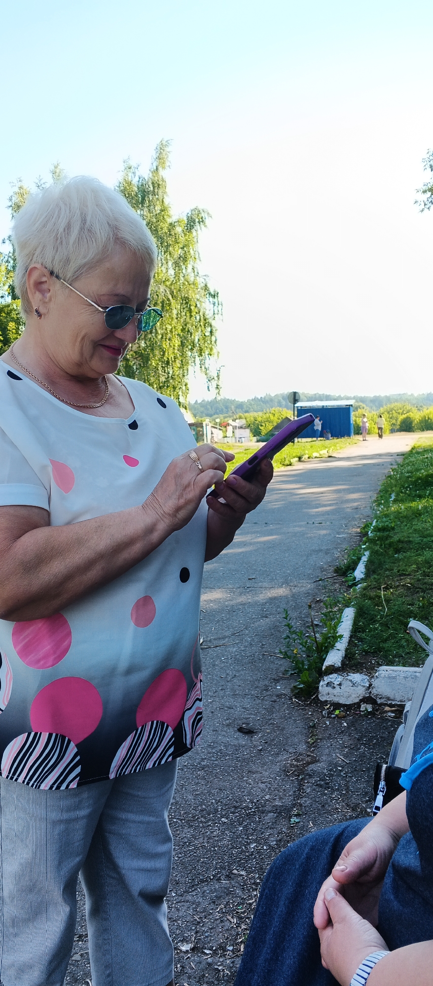 An elderly Caucasian woman with short blonde hair and sunglasses stands outdoors, holding a purple smartphone and looking at its screen. She wears a white blouse with pink and black circular patterns and gray pinstripe pants. She has a gold chain necklace and a ring on her right hand. The background shows a paved path, grass, trees, and a blue building in the distance. The scene appears to be a public area in the city of Podgornoye, Russia.